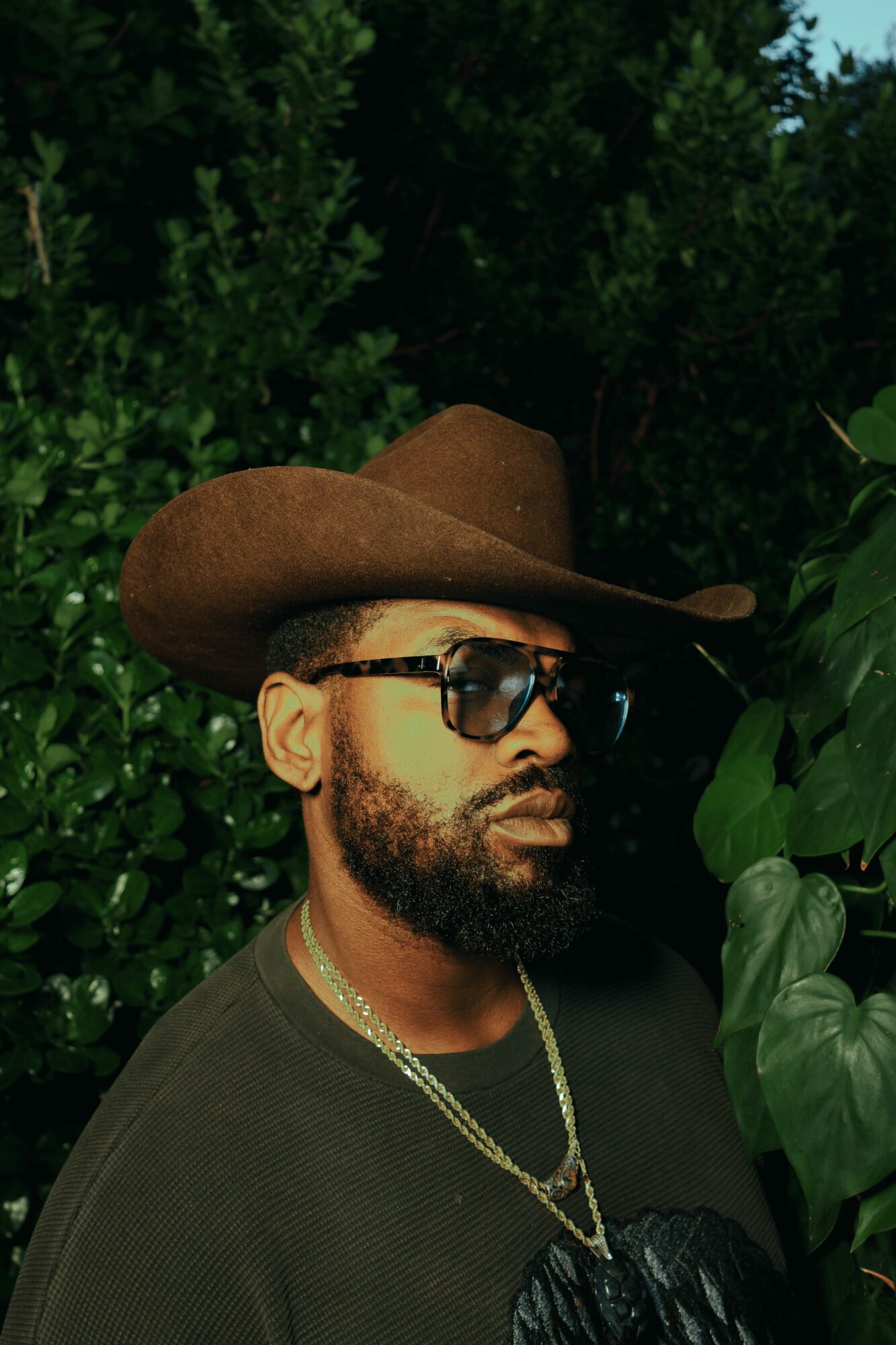 Man with beard wearing sunglasses and a wide-brimmed hat outdoors among green foliage.