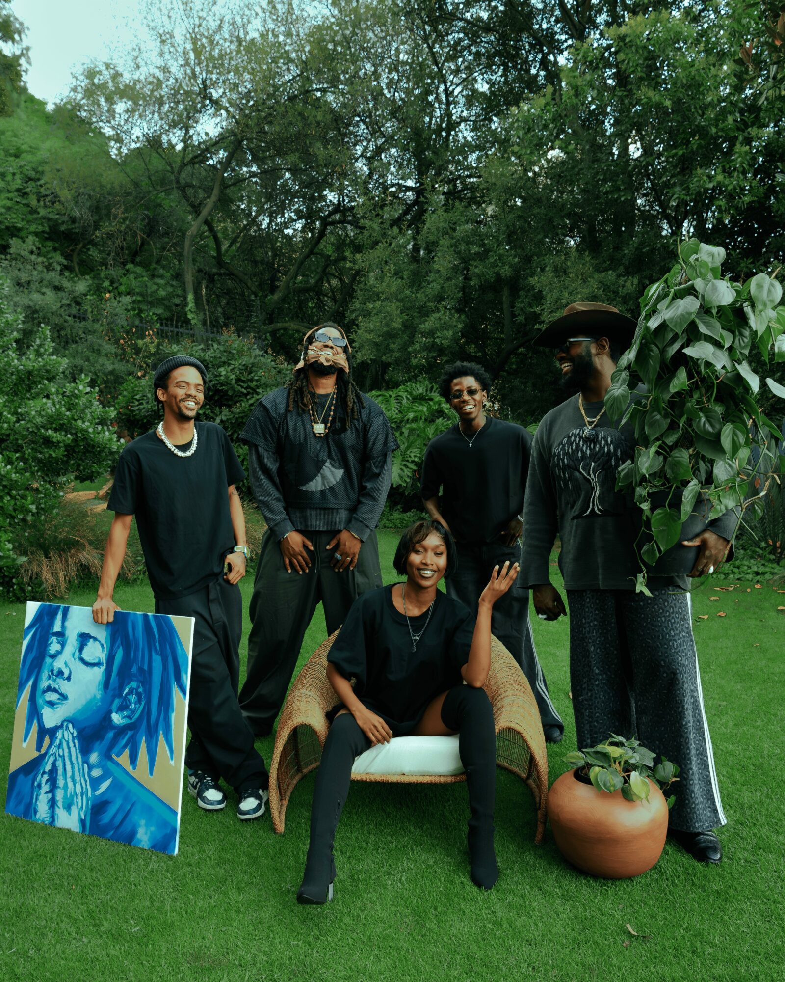 Five people outdoors, one sitting on a bench, others standing, with trees and greenery in background.