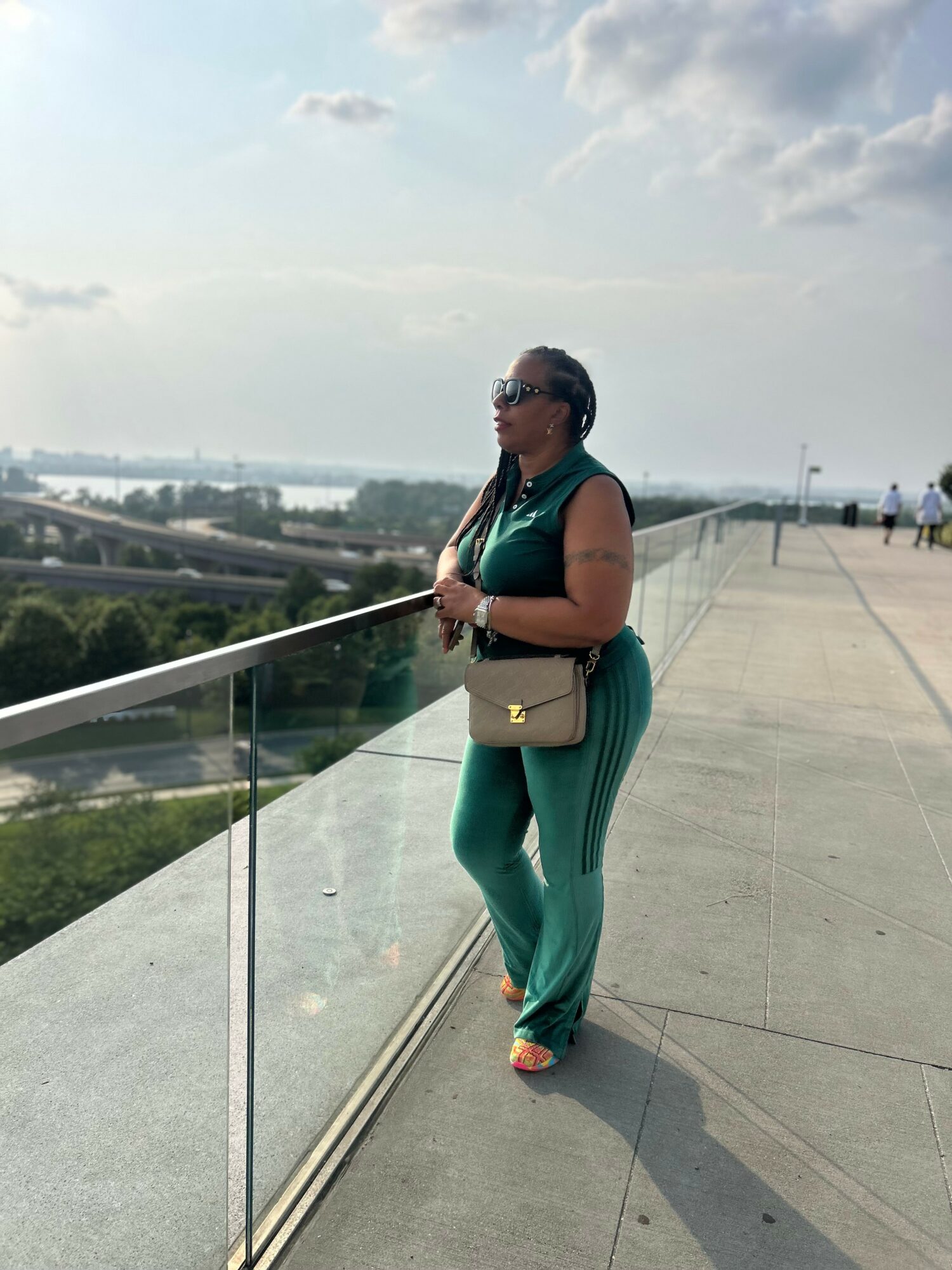 Woman standing on a balcony overlooking a cityscape with water, wearing sunglasses, a sleeveless top, and teal pants, holding a purse.