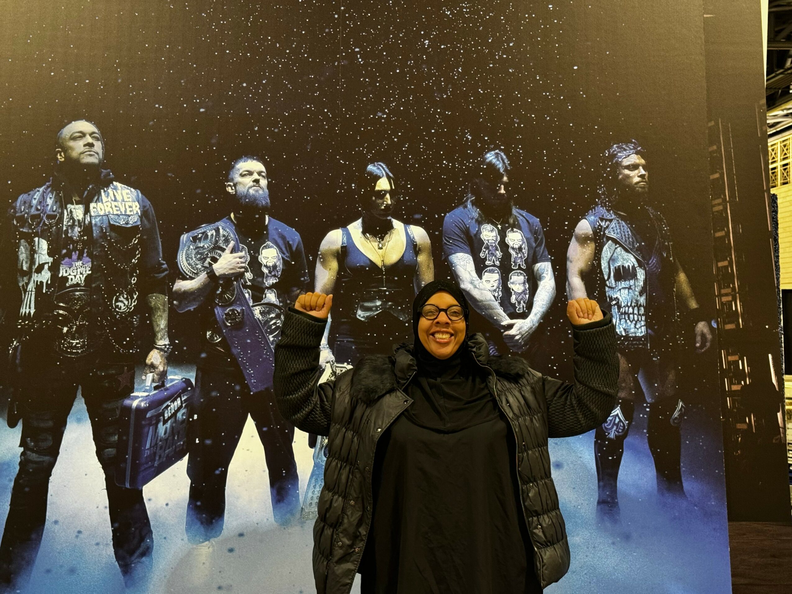 Person smiling and wearing glasses, raising fists, standing in front of a large poster of five people in dark clothing, with a starry background.