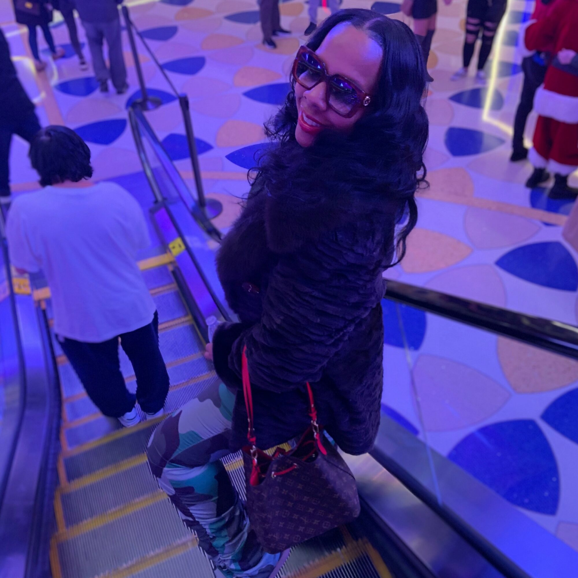 Woman with long dark hair and glasses smiling, standing on an escalator in a colorful indoor setting.