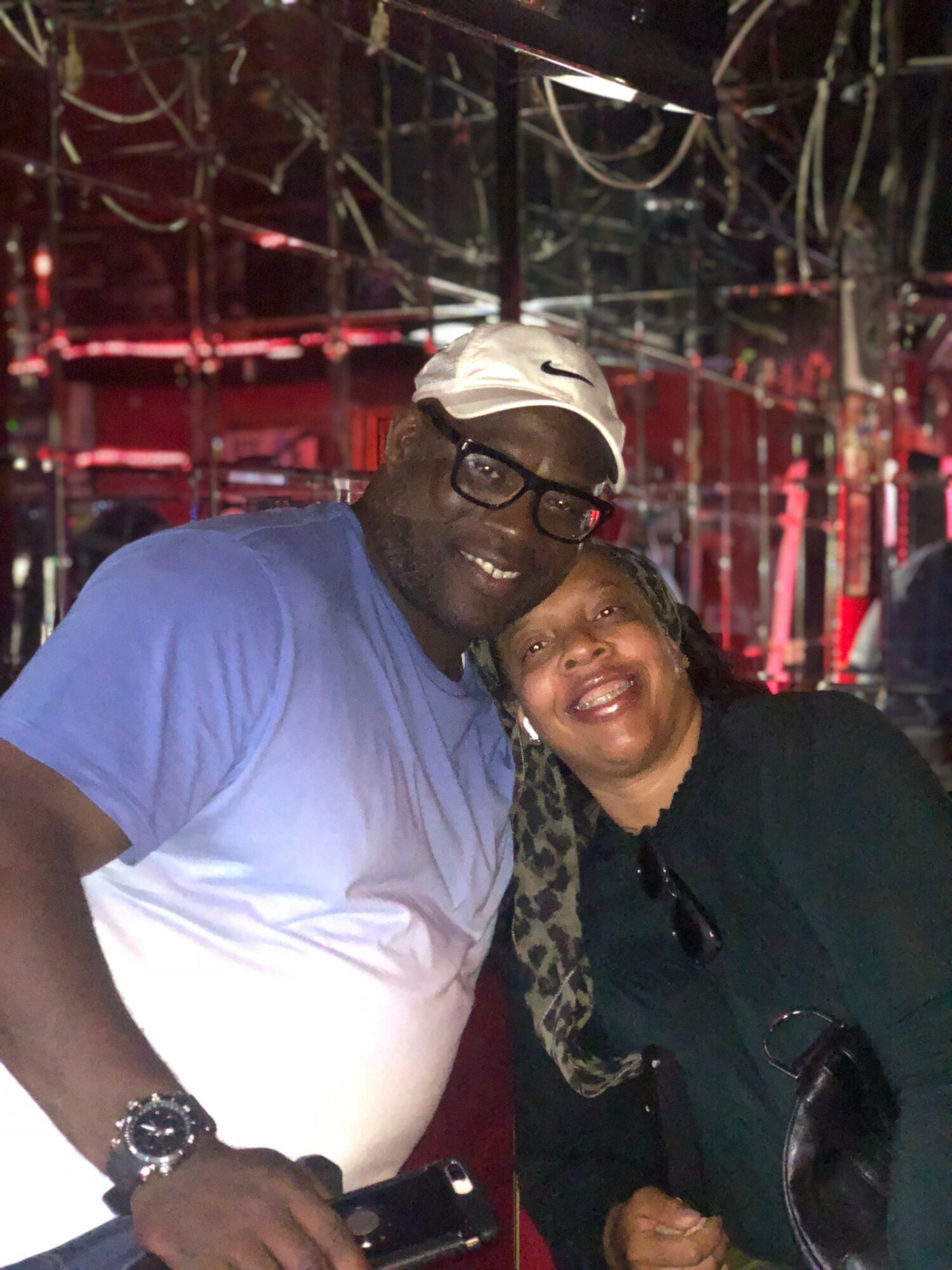 Two people smiling, one wearing glasses and a white cap, the other with curly hair, in a dimly lit environment with red lighting.