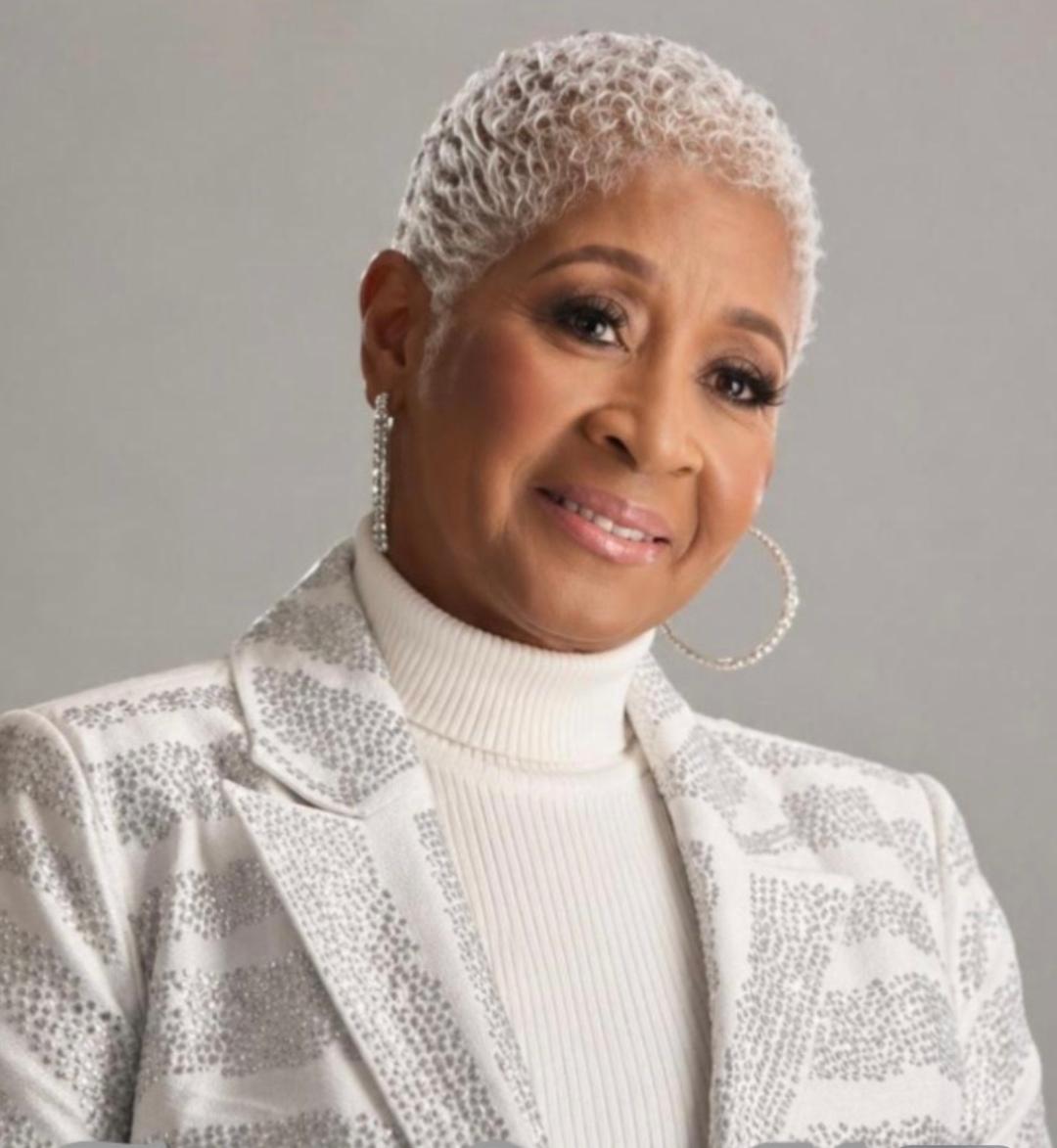 A woman with short, curly gray hair wearing a white turtleneck and patterned blazer, smiling softly, against a plain background.