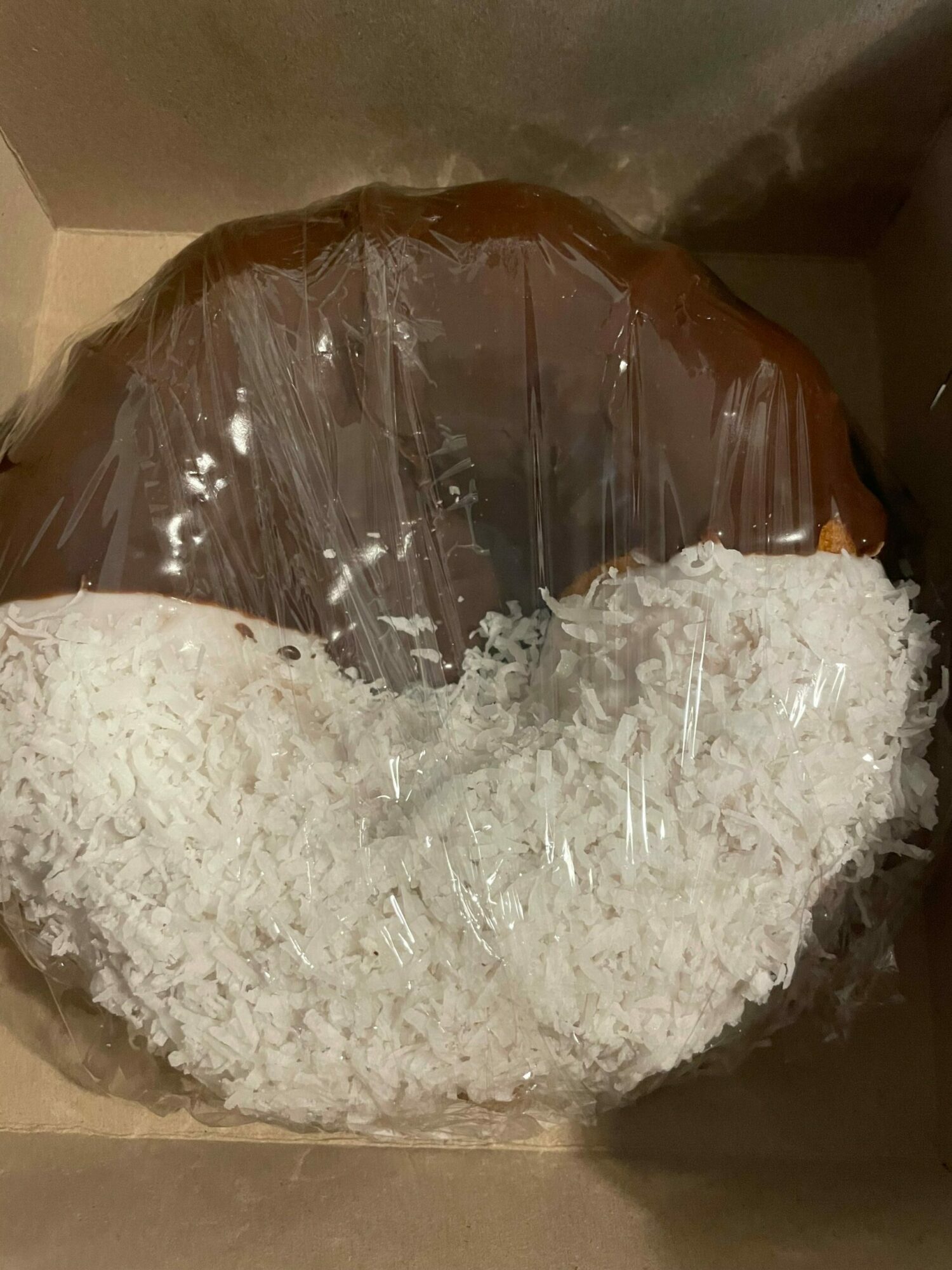 Chocolate cake with white shredded topping inside a box, partially covered with plastic wrap.