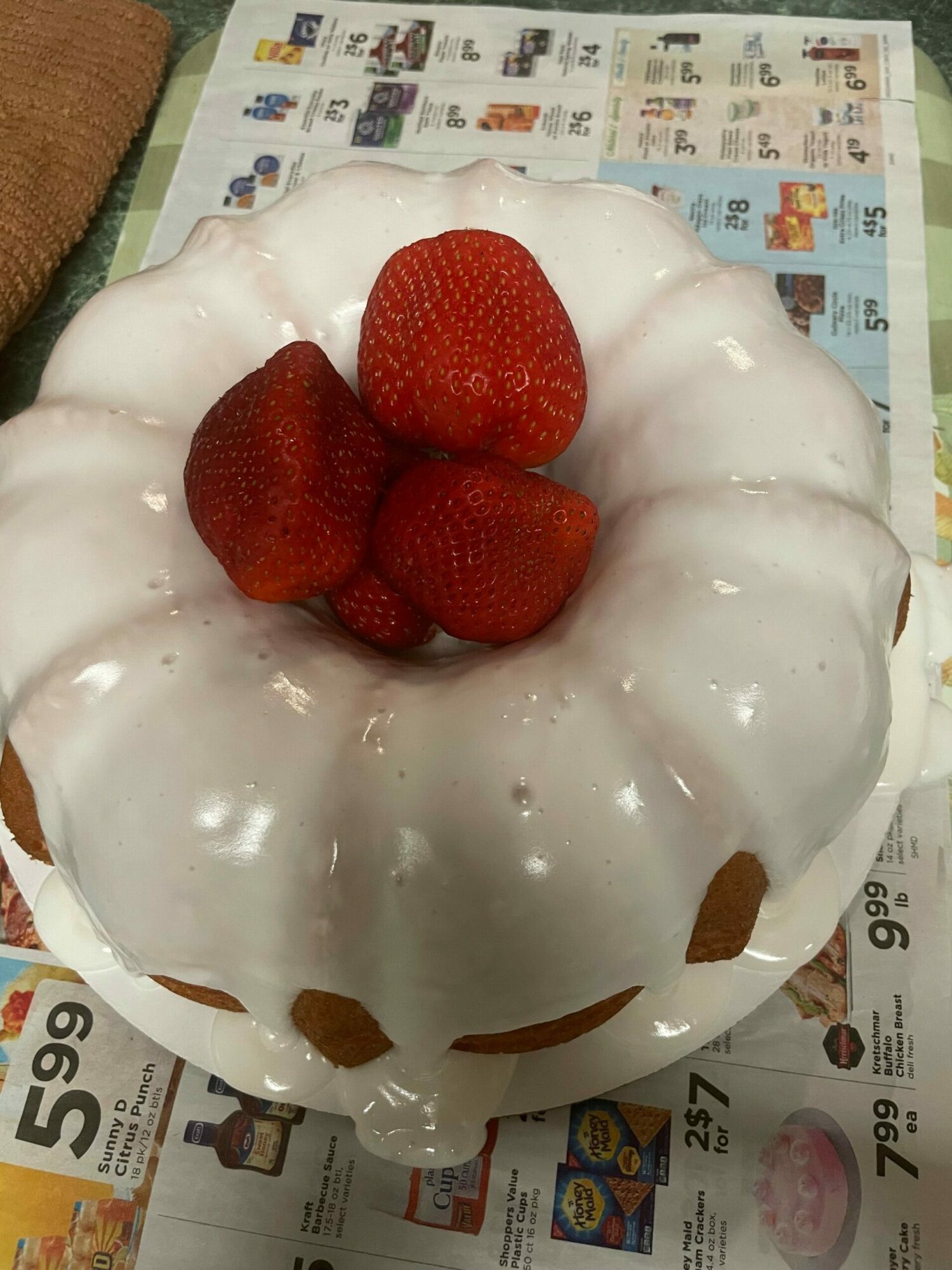 A bundt cake with white icing and strawberries on top, placed on newspaper. The cake has brown spots and a hole in the center.