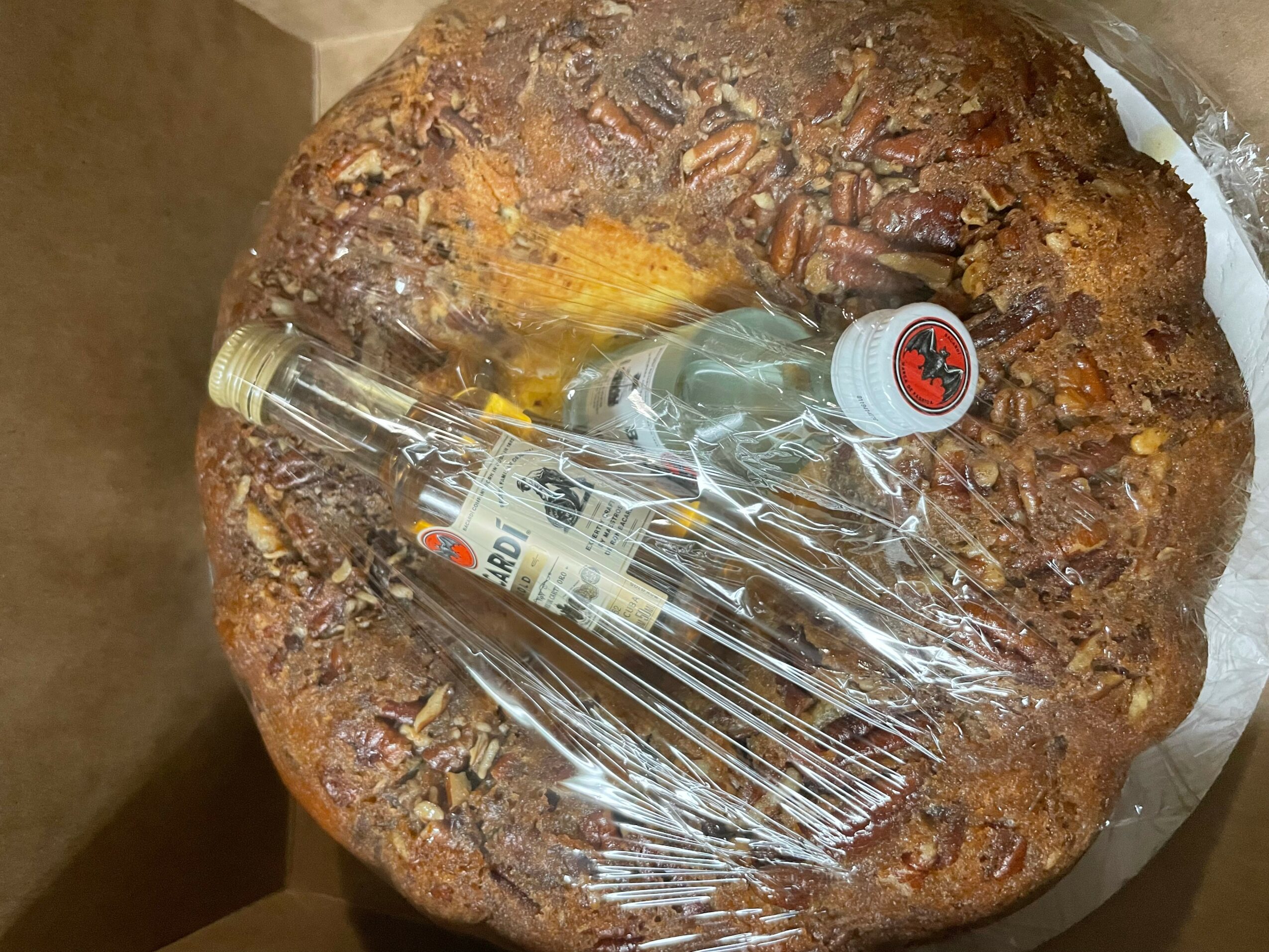 Large round baked item with a bottle and a small container on top, wrapped in plastic, inside a box.