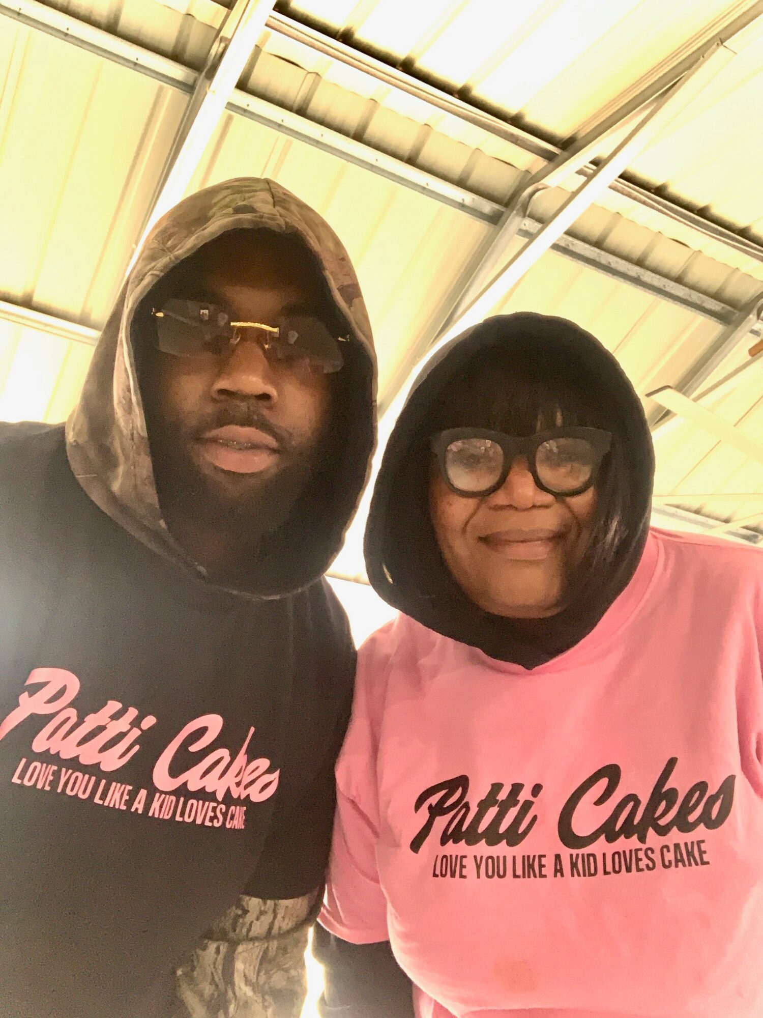 Two people wearing pink shirts with Patti Cakes logo, standing under a metal roof, smiling at the camera.