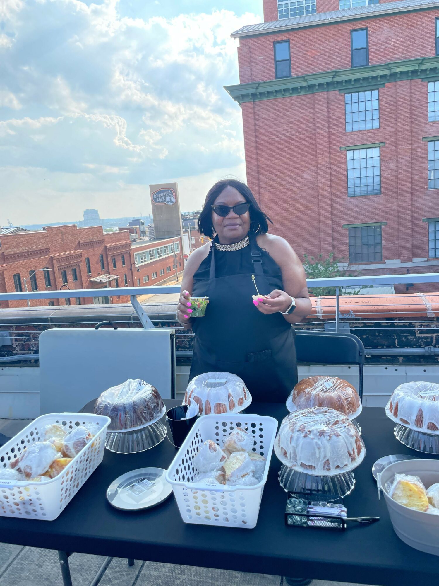 Woman with dark hair and glasses standing behind a table with baked goods, on a balcony with city buildings in background.
