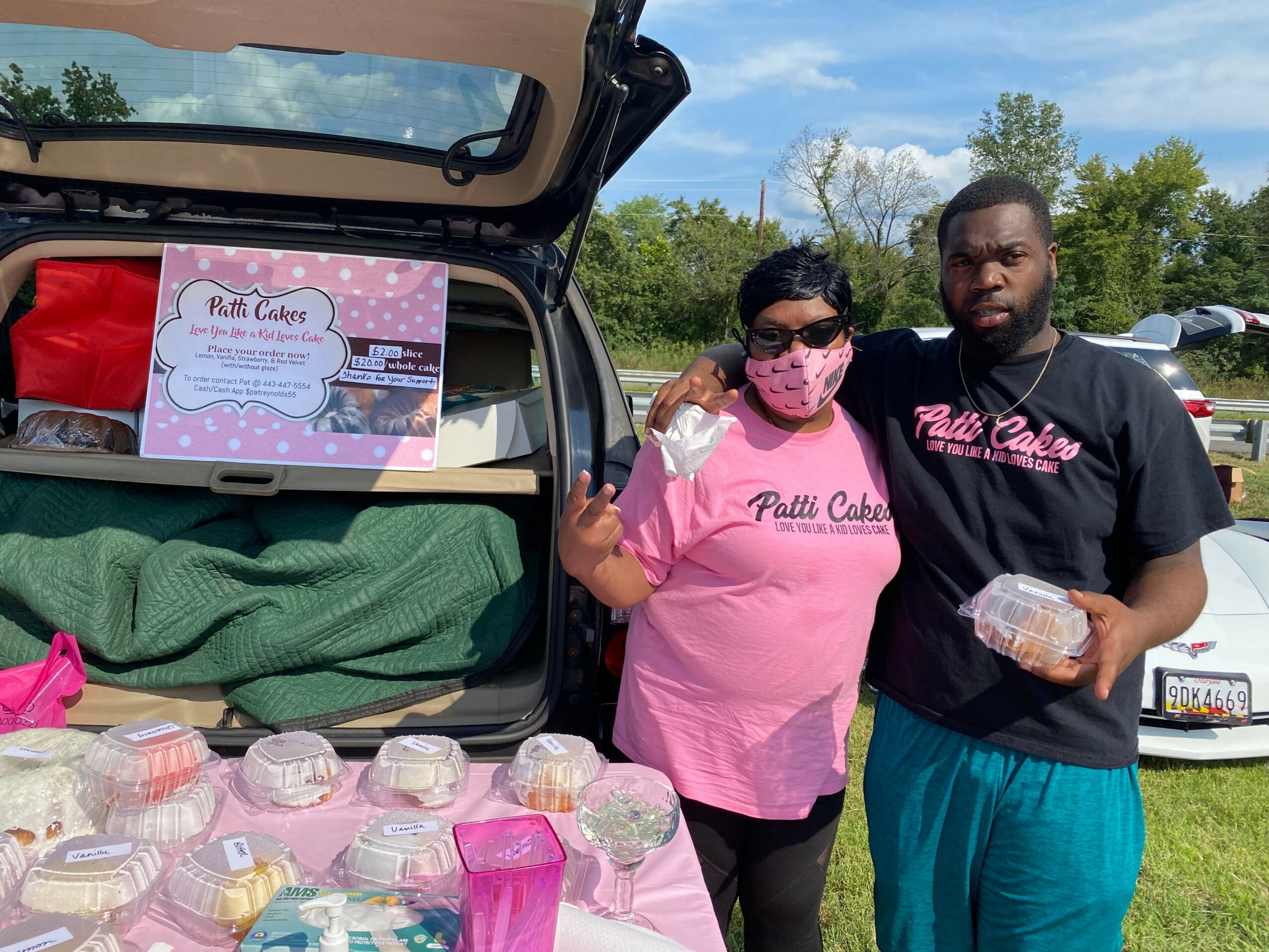 Two people stand outdoors next to an open car trunk filled with baked goods, with a sign inside the trunk. One person wears a pink shirt and mask, the other a black shirt, both holding containers of baked goods.
