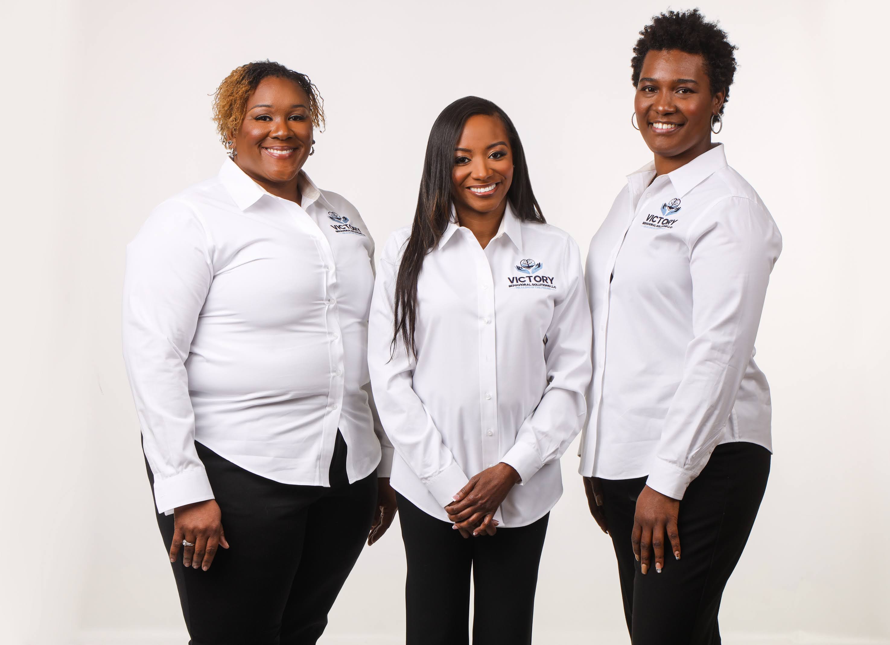 Three women in white shirts and black pants standing together smiling against a white background.
