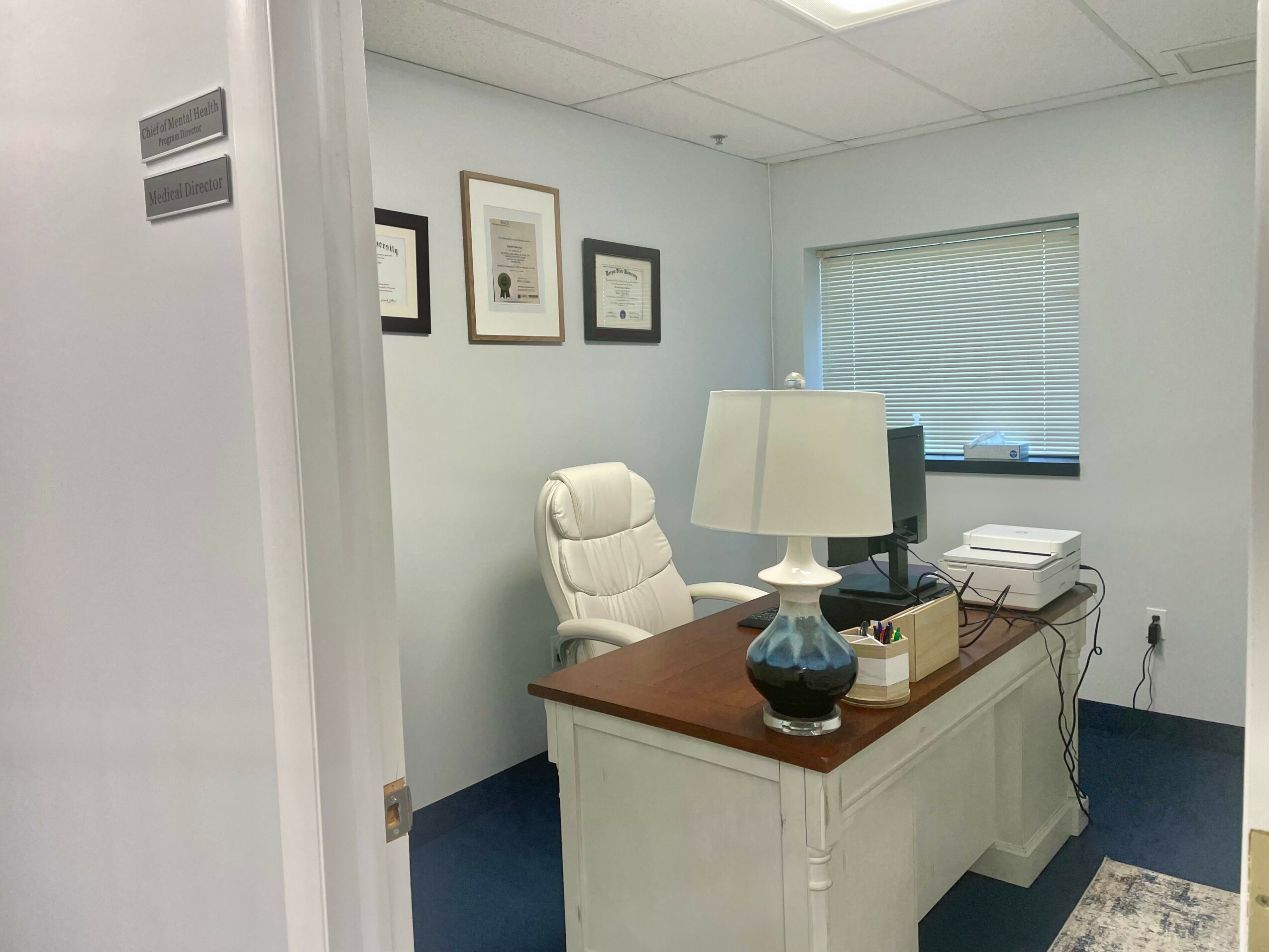 Office with white walls, a window with blinds, framed pictures, a desk with a lamp, computer, and printer, and a white chair.