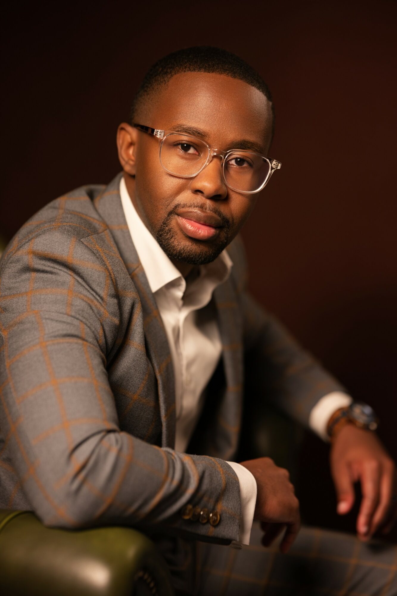 Portrait of a man wearing glasses and a gray suit, sitting with hands on a table, dark background.