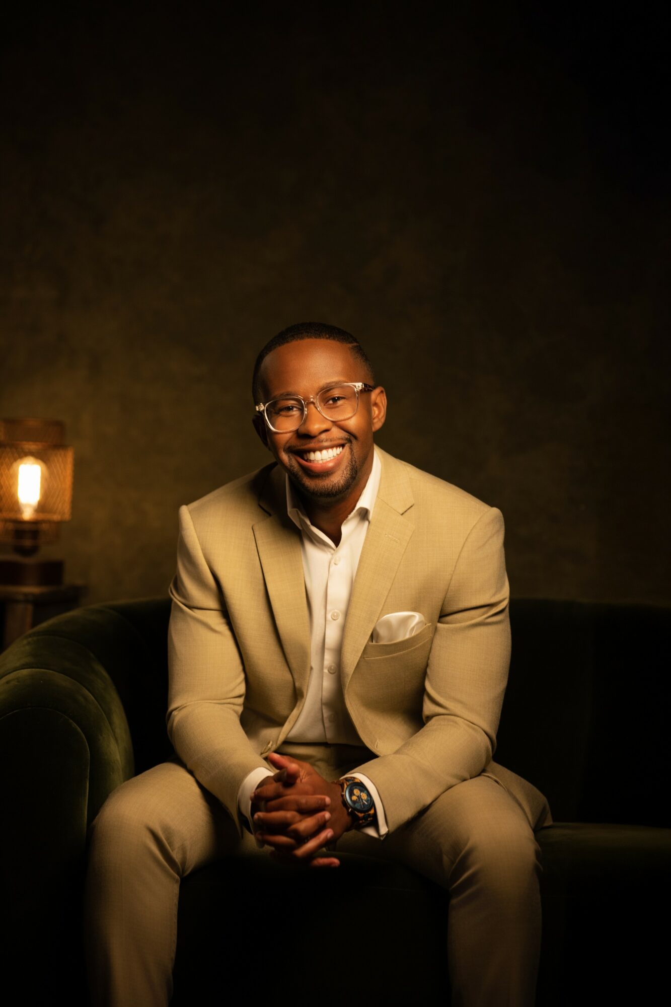 Man in beige suit and glasses sitting on a dark sofa, smiling, with warm lighting in background.