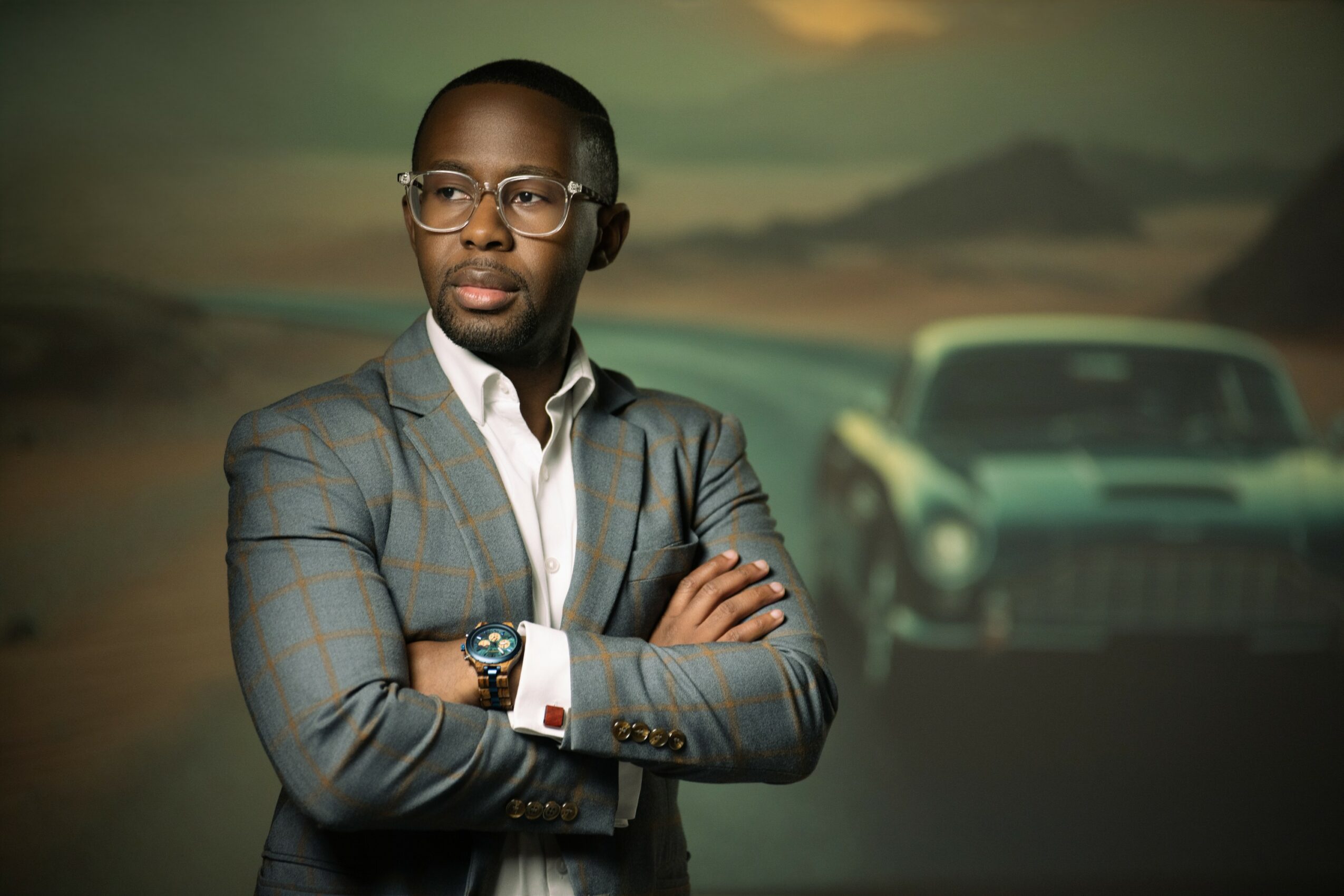 Man in glasses and suit with arms crossed, standing in front of a vintage car and landscape background.