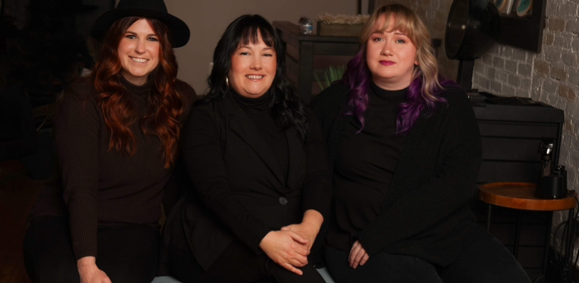 Three women sitting together indoors, smiling, with dark clothing and a brick wall in the background.