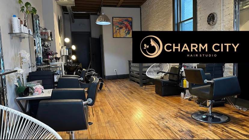 Interior of a hair salon with chairs, mirrors, and wooden flooring, featuring a Charm City Hair Studio sign on the wall.