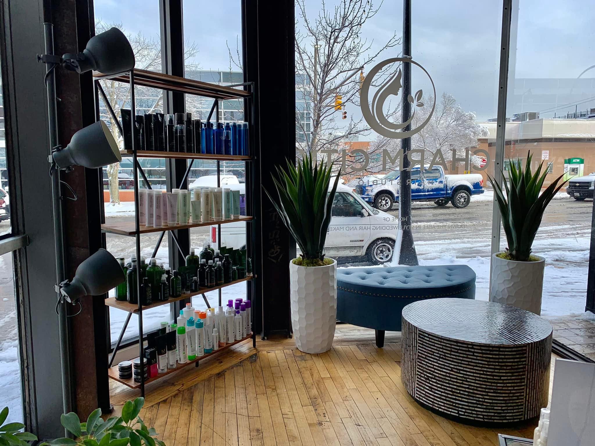 Indoor space with large windows, potted plants, a round ottoman, and a shelving unit with nail polish bottles.