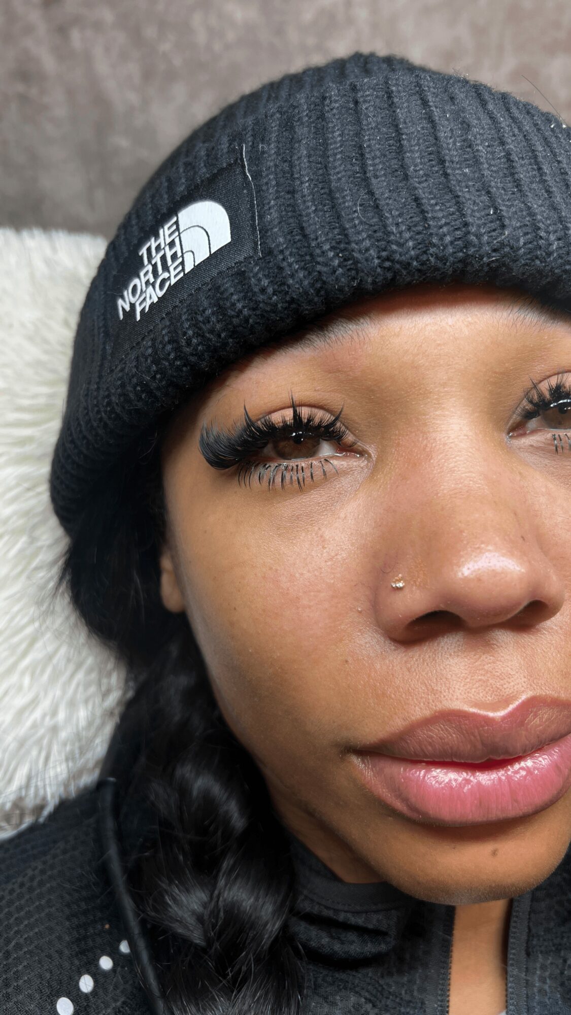 Close-up of a woman wearing a black beanie with North Face logo, showing her face with makeup and a nose piercing.