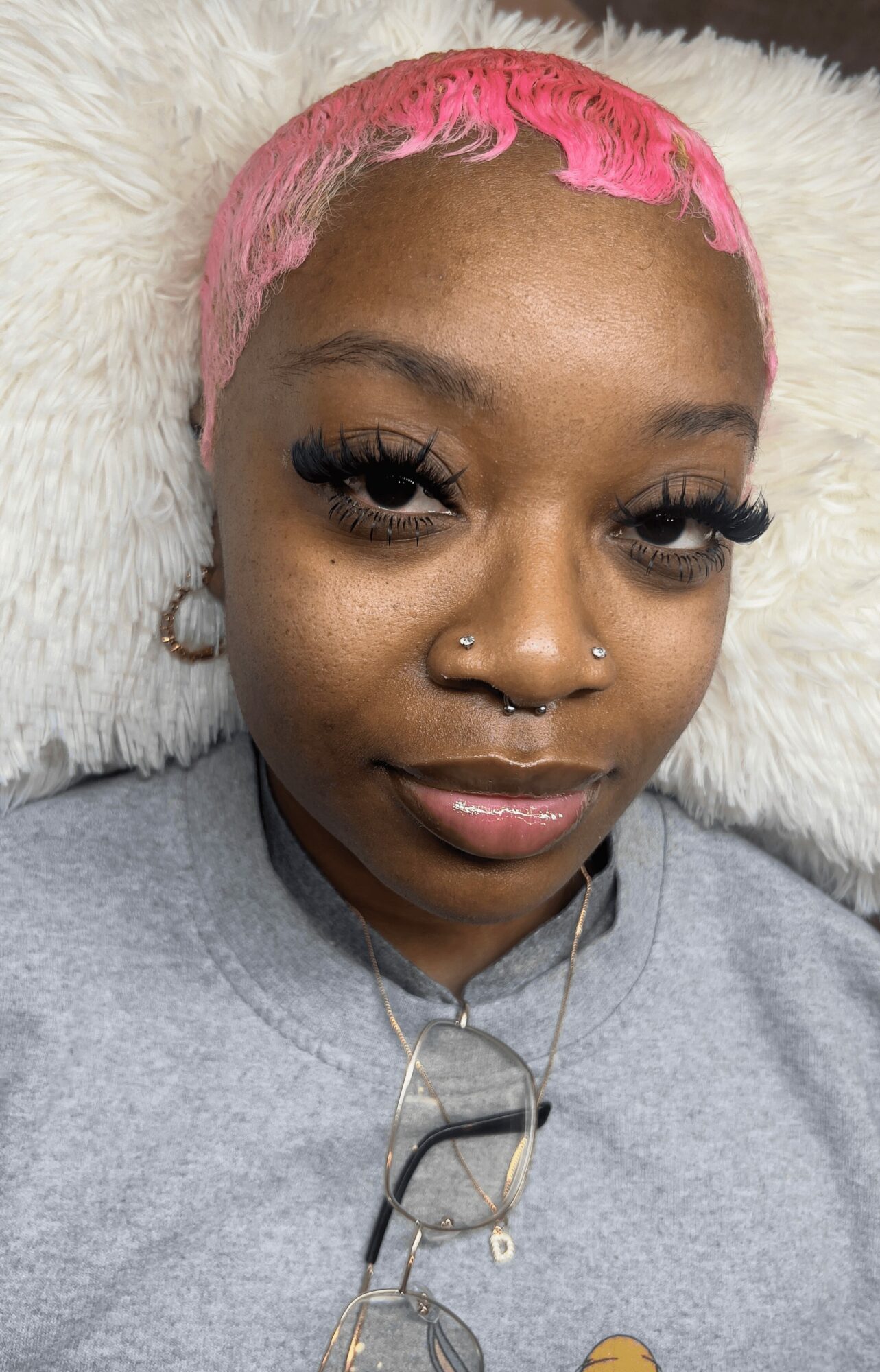 Young woman with short pink hair, multiple facial piercings, and long eyelashes, lying on a white fluffy surface.