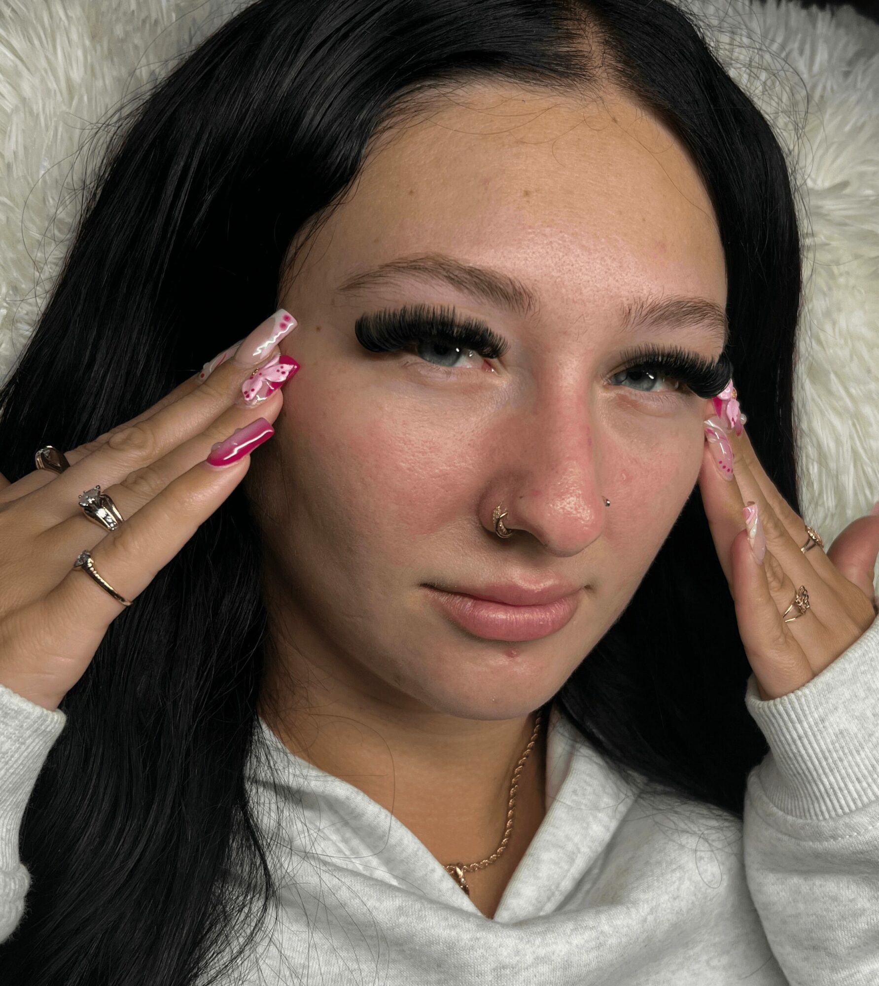 Close-up of a woman with long dark hair, makeup, and jewelry, touching her face with both hands, wearing a white hoodie.