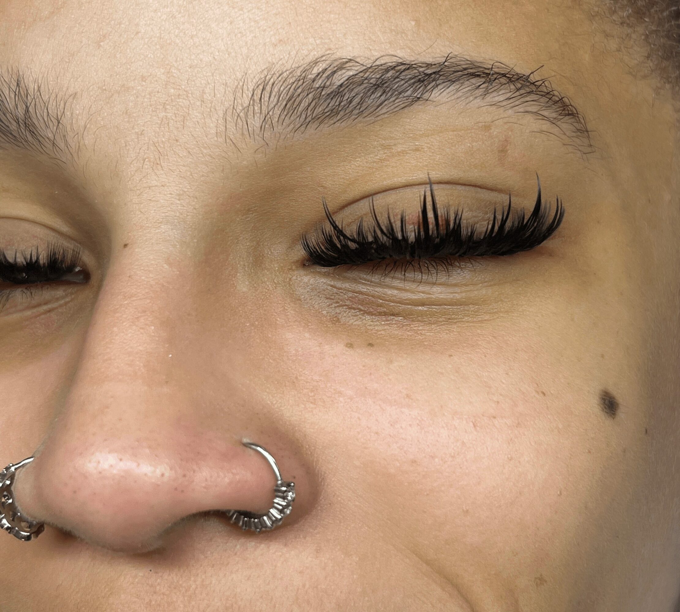 Close-up of a person's face with closed eyes, showing long eyelashes, a nose piercing, and a small mole on the cheek.