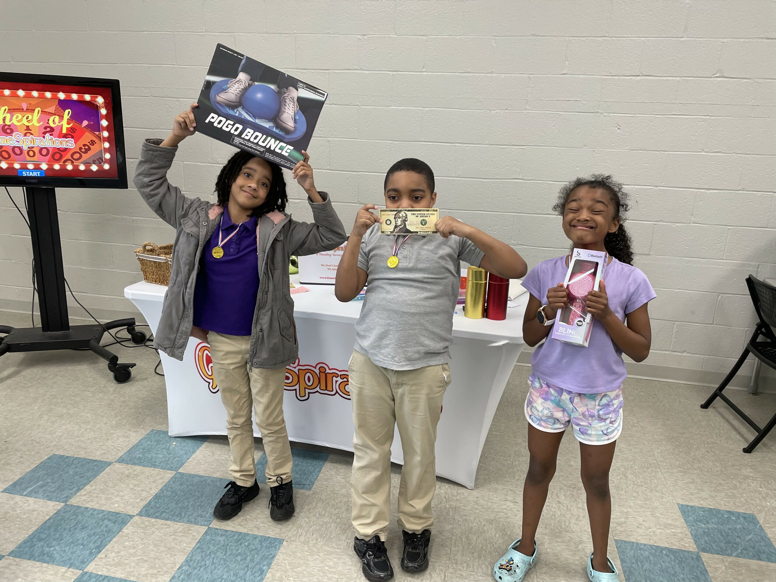 Three children stand in front of a table, holding signs and smiling, in an indoor setting with a white brick wall.
