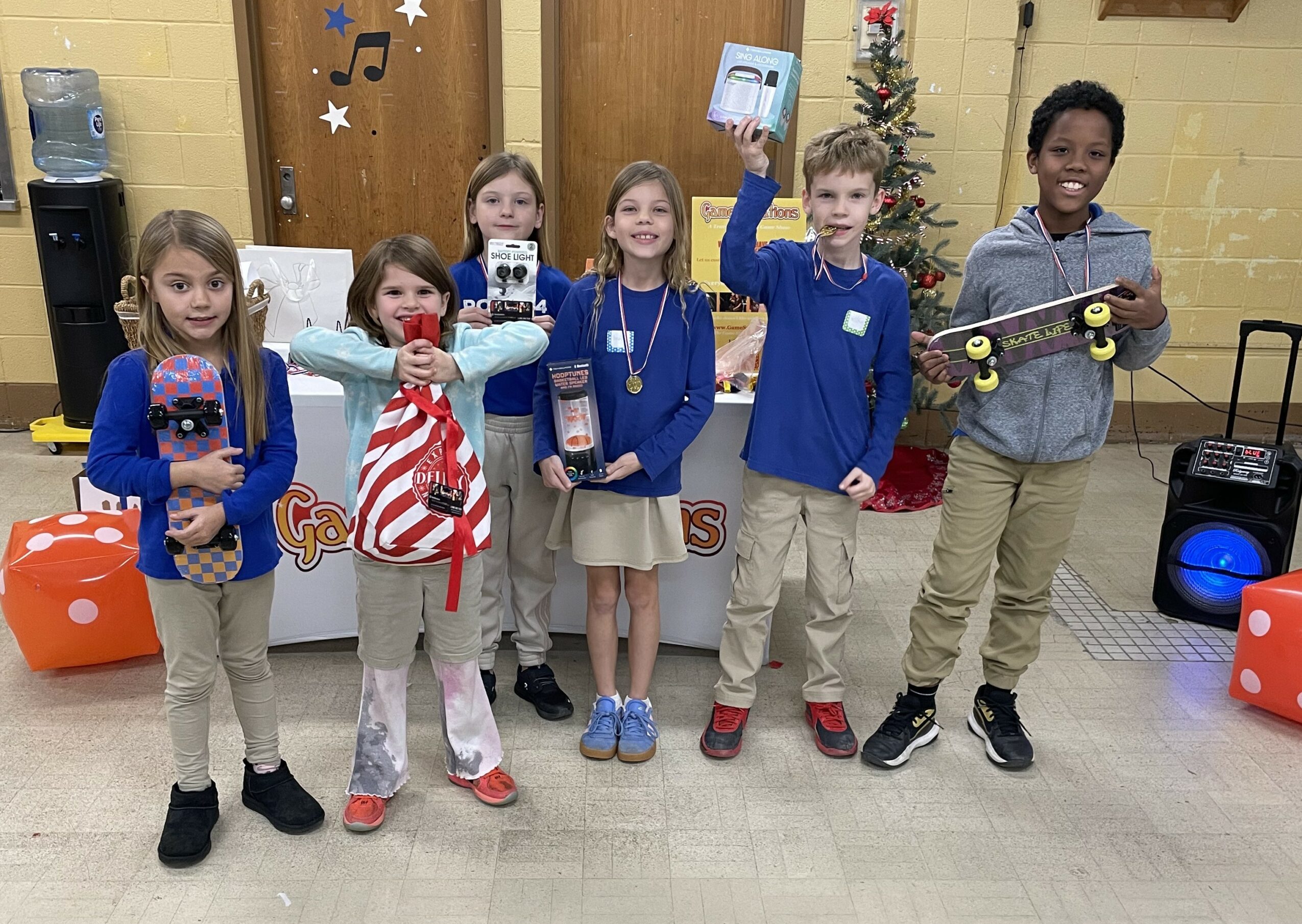 Six children stand indoors holding skateboards and gifts, smiling, with a Christmas tree and decorations in the background.