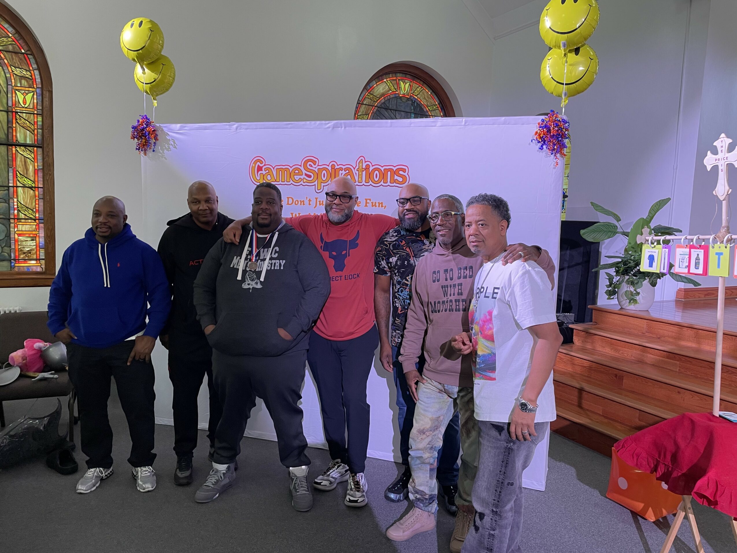 Group of six men standing inside a church with balloons and a banner, smiling for a photo.