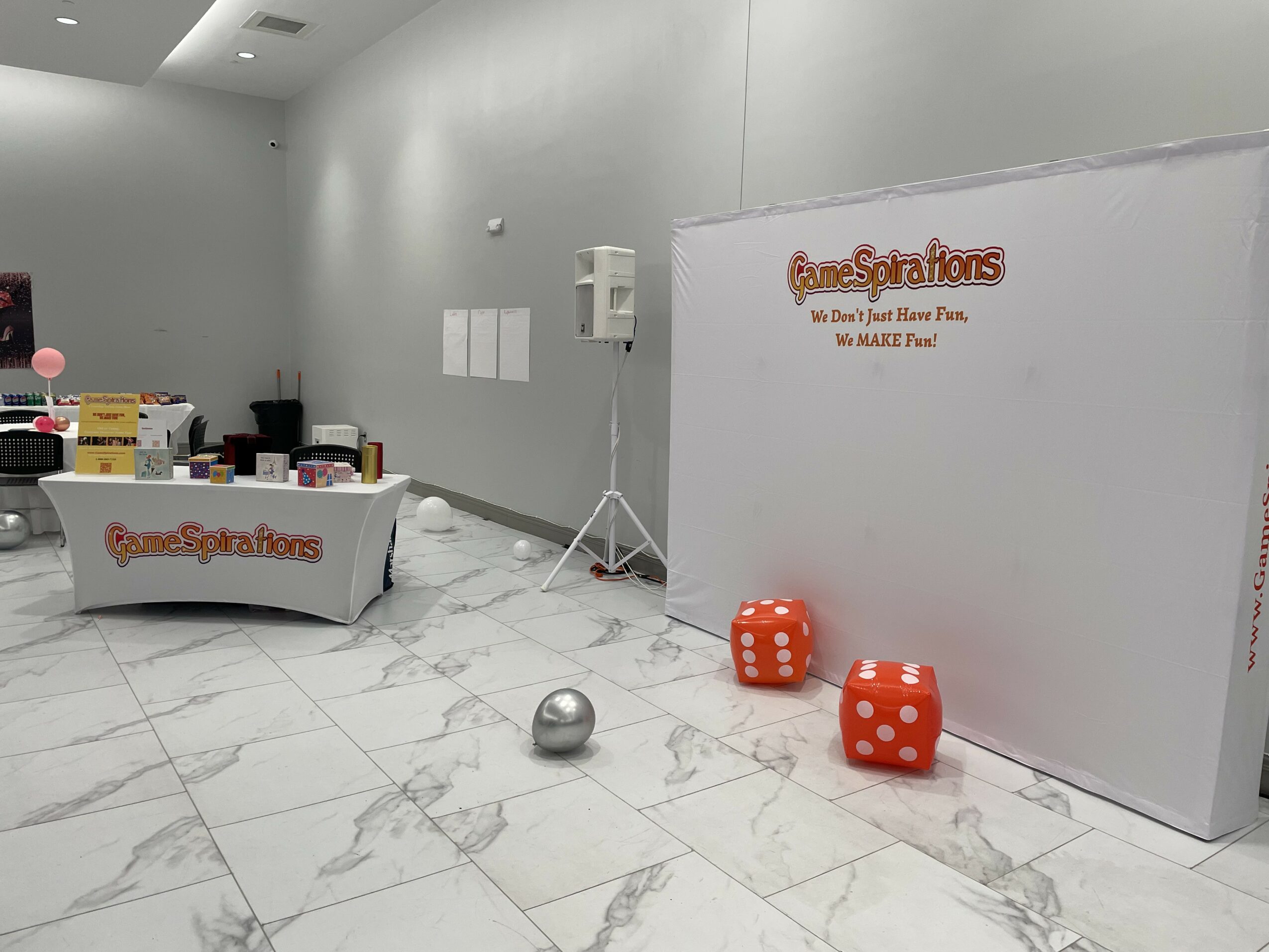 Room with a table, boxes, and large foam dice, white walls, and marble floor, with a white backdrop and some equipment.