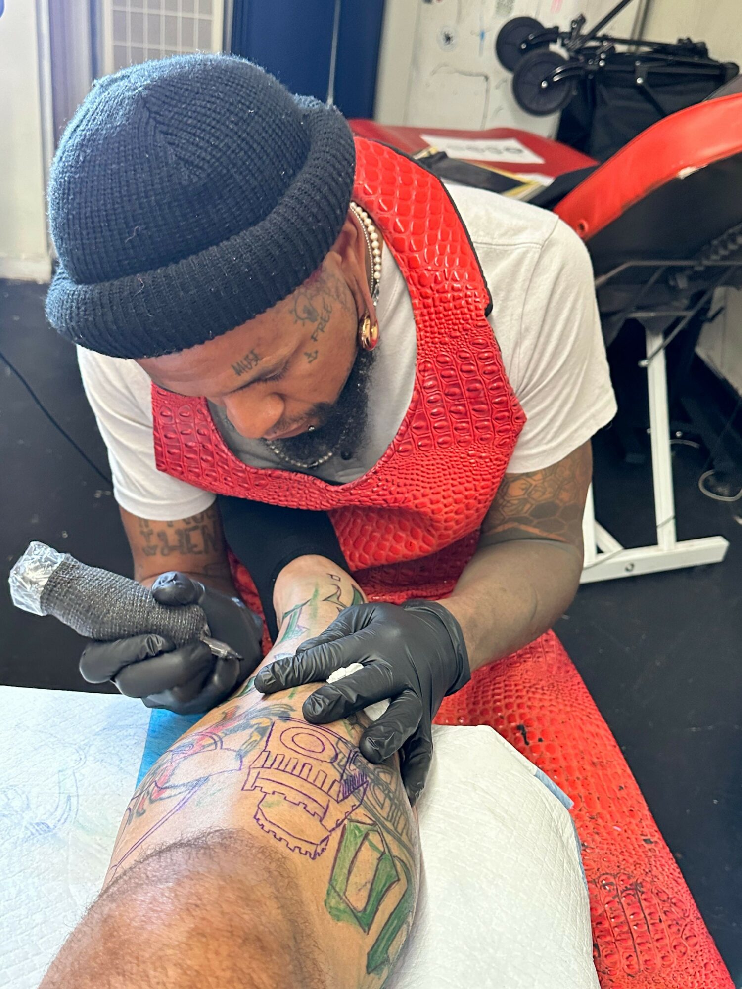 Tattoo artist working on a client's arm, wearing gloves and a red vest, with a tattoo machine in hand.
