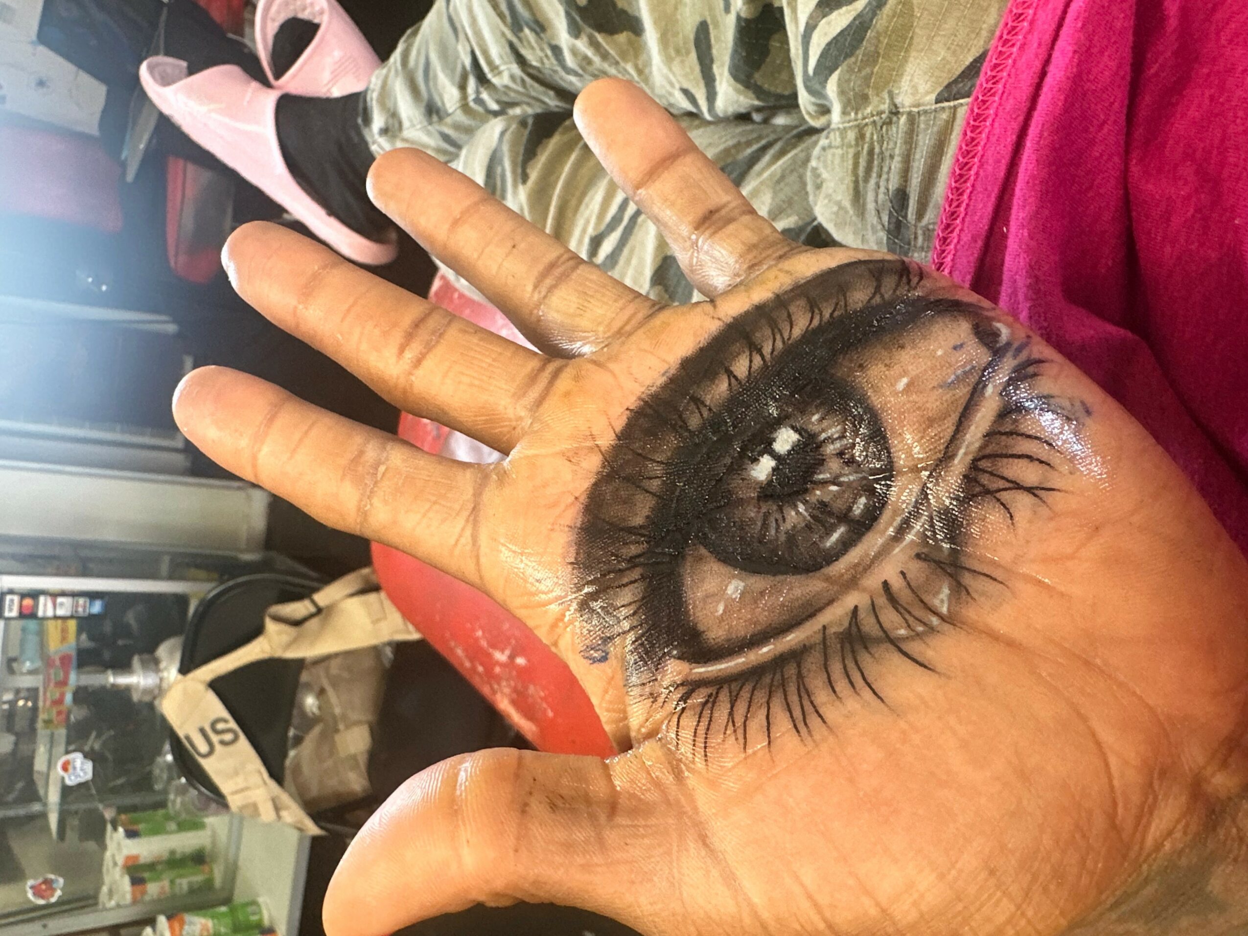 A hand with detailed eye and eyelashes painted on the palm, held up against a background of a person lying down.