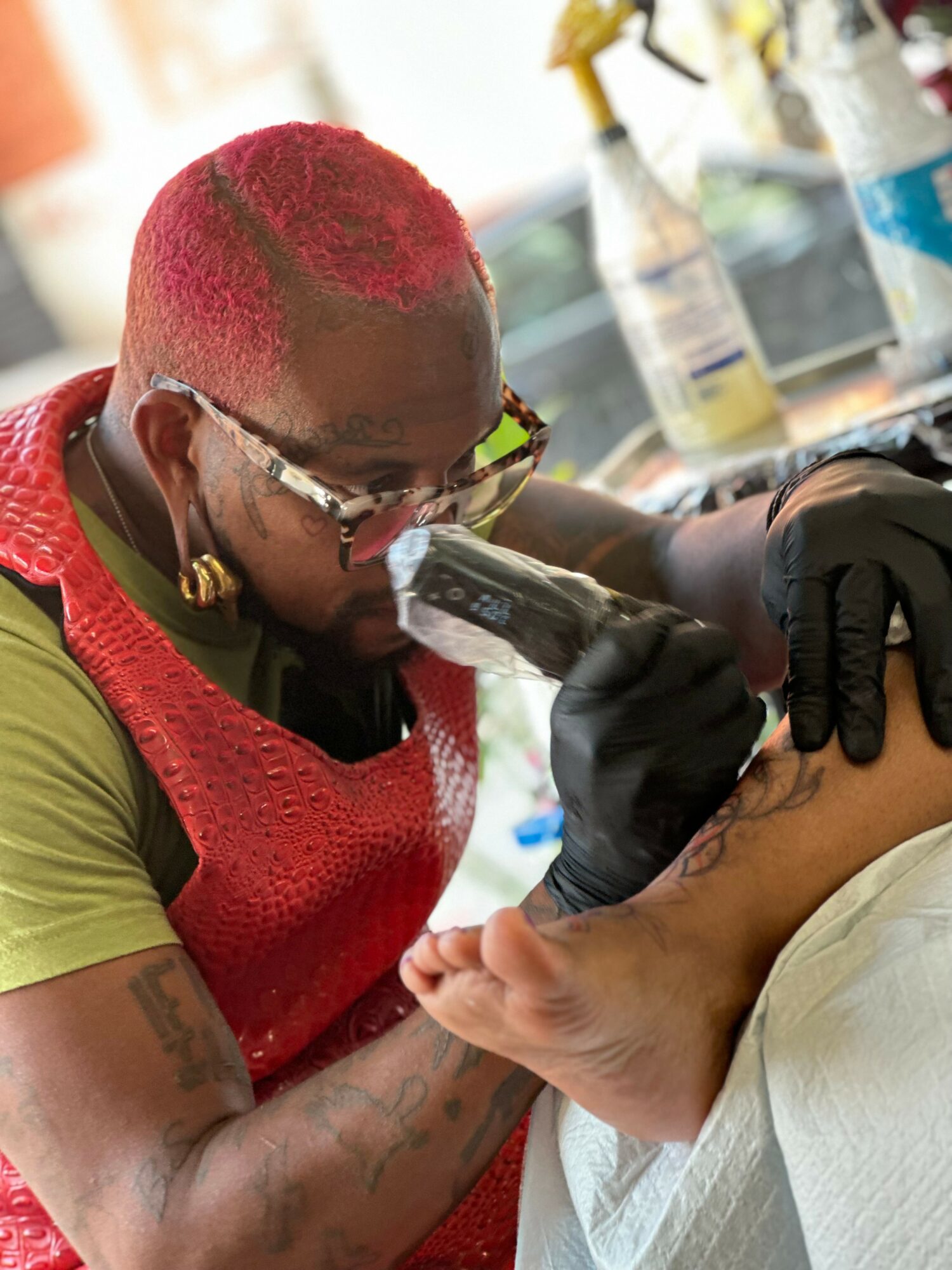 Tattooed person with pink hair getting a tattoo on their foot by an artist wearing gloves.