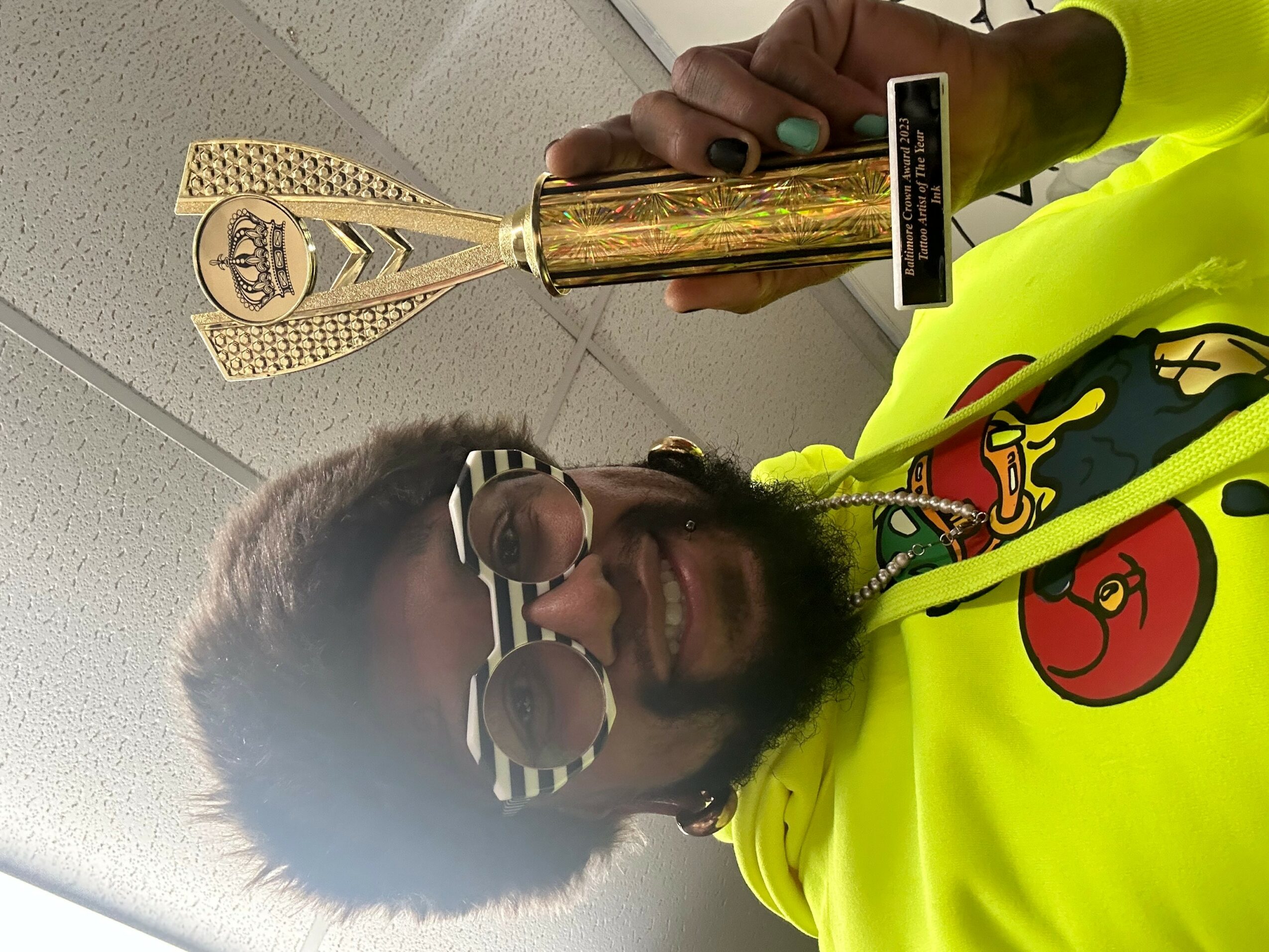Person with sunglasses and a beard holding a trophy, wearing a bright yellow hoodie with a cartoon character, smiling.