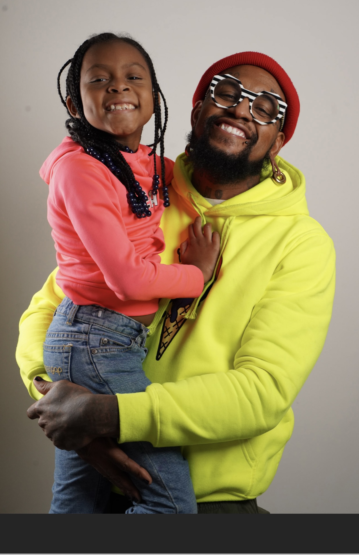 Smiling man holding girl, both wearing colorful clothing, against a plain background.