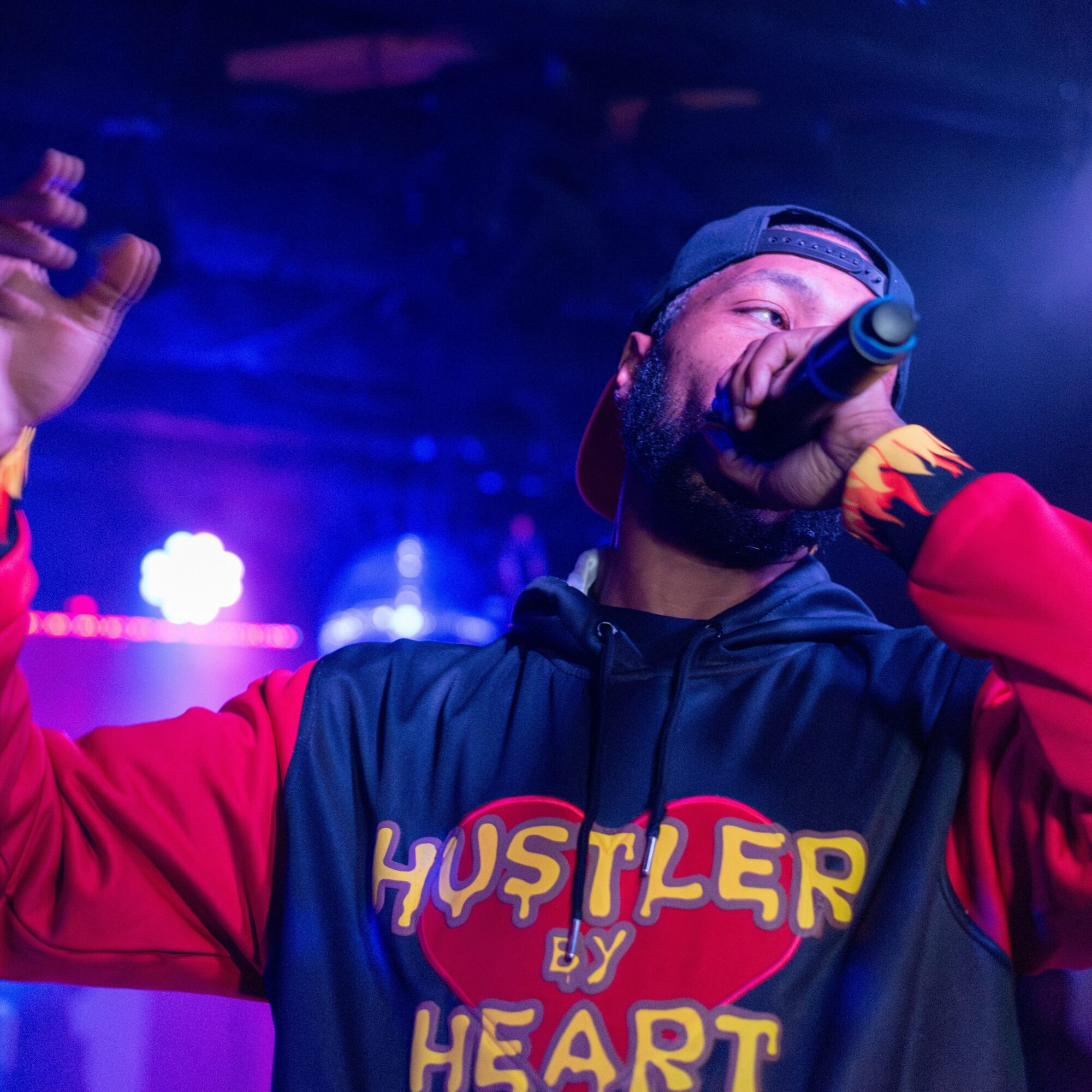 Performer singing into microphone on stage, wearing hoodie with 'Hustler by Heart' logo, cap backwards, with stage lights in background.