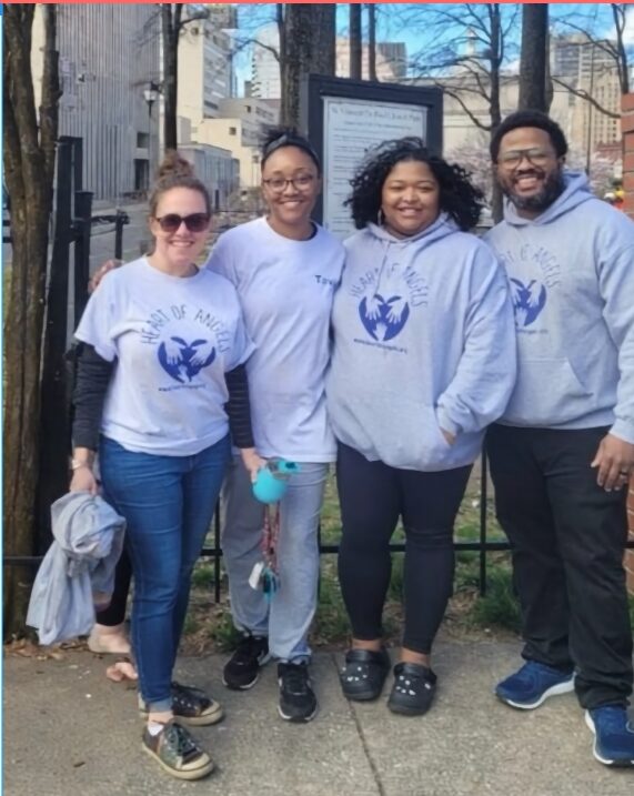 Four people standing outdoors, wearing matching light gray sweatshirts with a blue logo, smiling, with trees and a fence in the background.