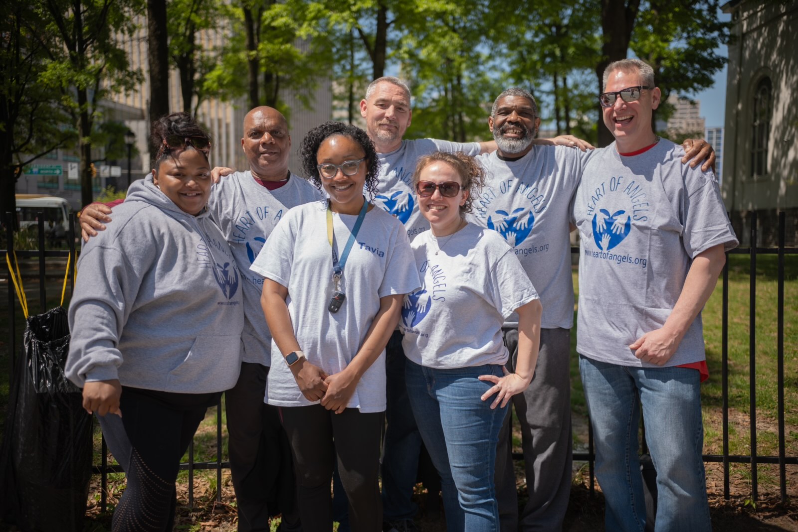 Group of seven people outdoors, smiling, wearing matching gray T-shirts with a blue logo, standing in front of trees and a fence.