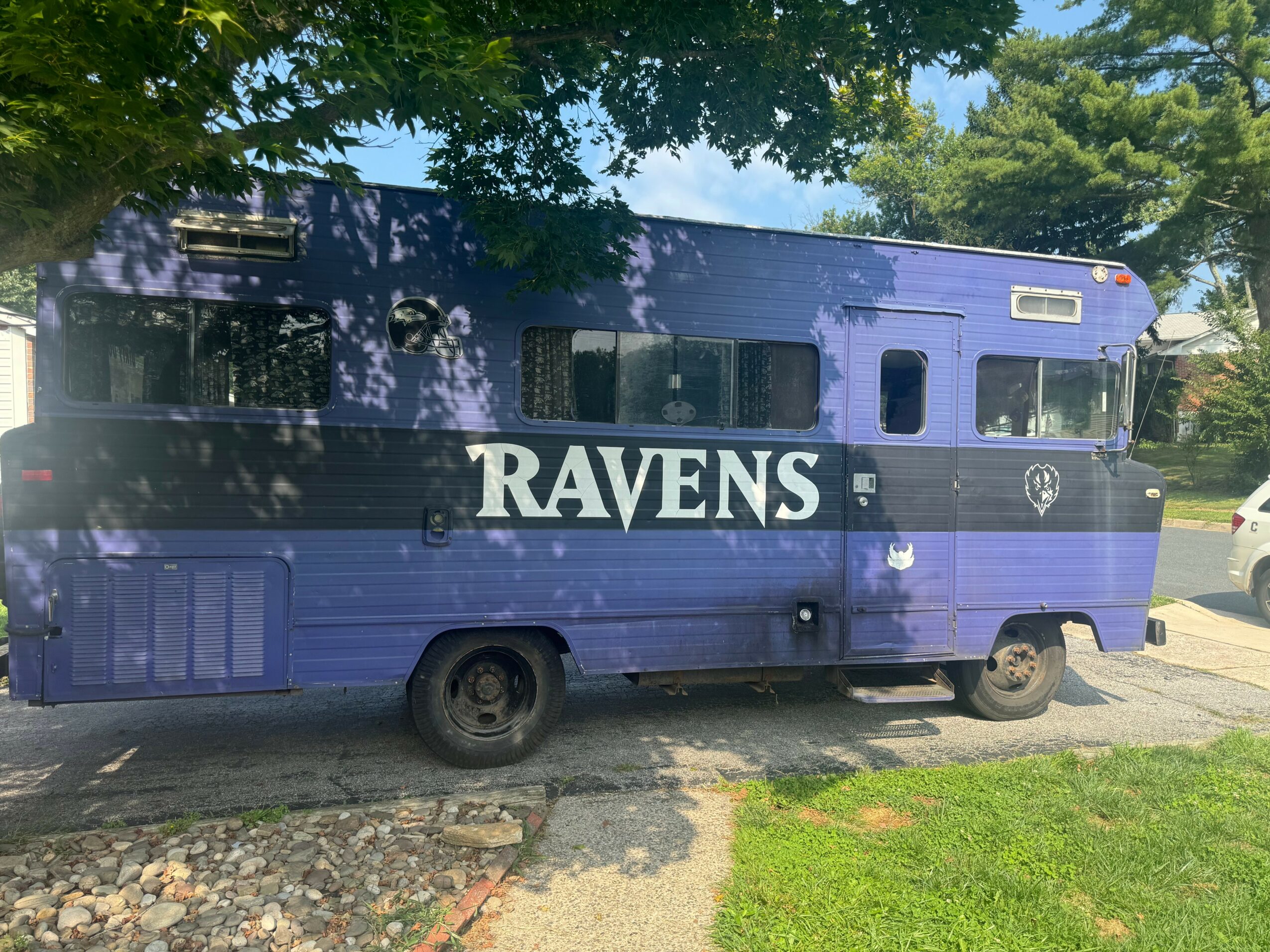 Blue RV with white text 'RAVENS' parked on street, trees and grass in background.
