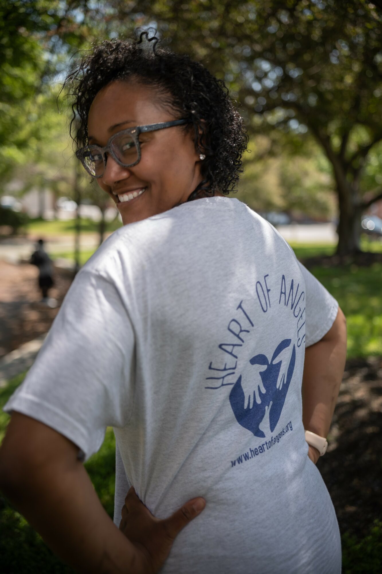 Smiling woman with glasses outdoors, wearing a white T-shirt with a blue logo and text on the back, trees in background.
