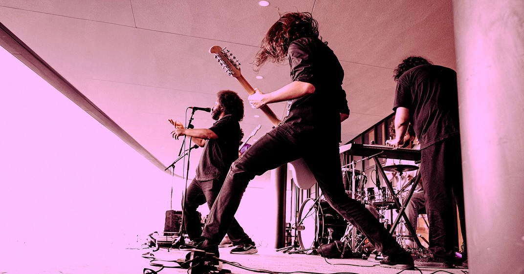 Band performing on stage with musicians playing guitars and singing, stage lighting creates a pinkish hue.
