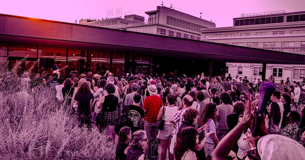 Large crowd of people gathered outside a modern building with glass windows, some raising their hands, in an outdoor setting.