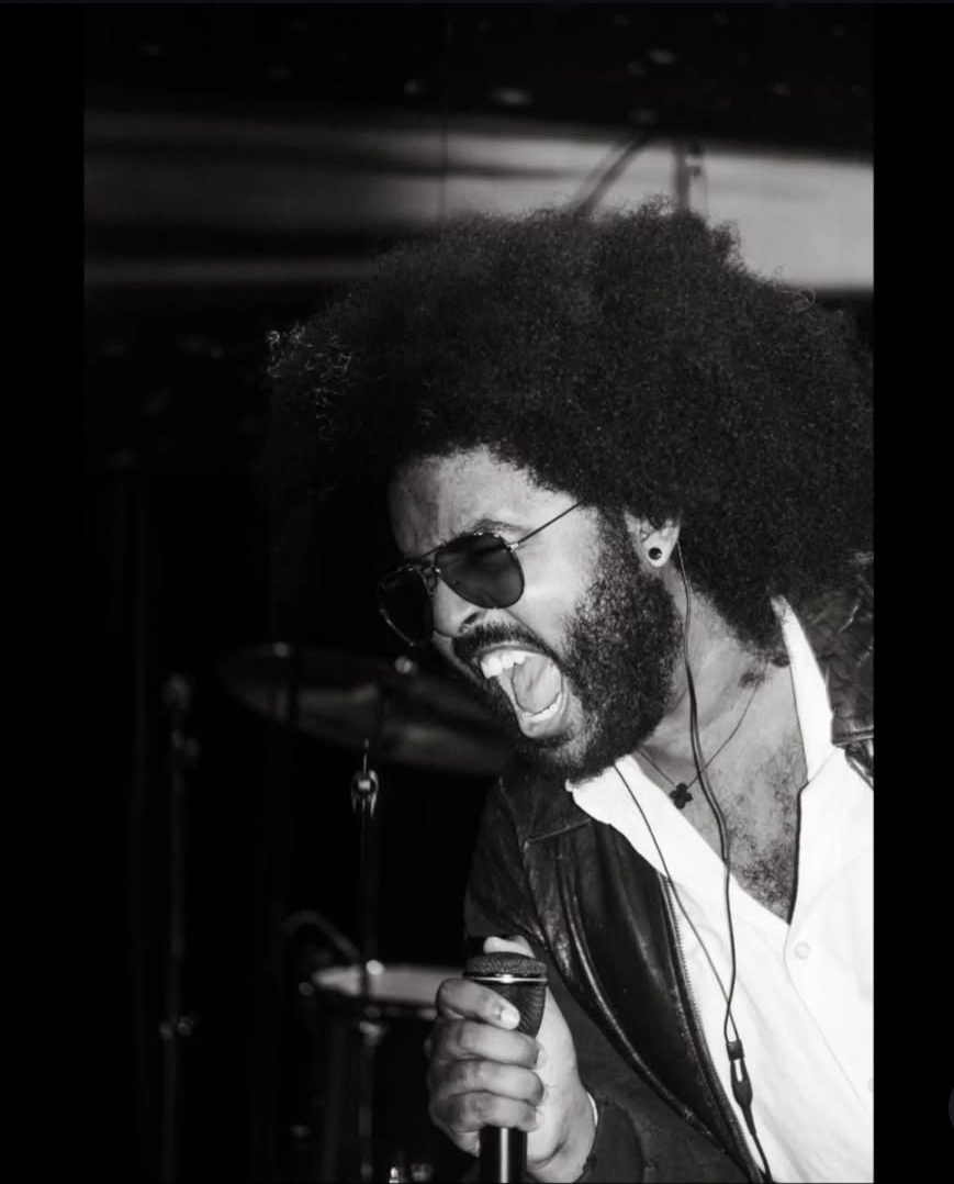 Person singing passionately with sunglasses, curly hair, and a leather jacket, holding a microphone, in black and white.