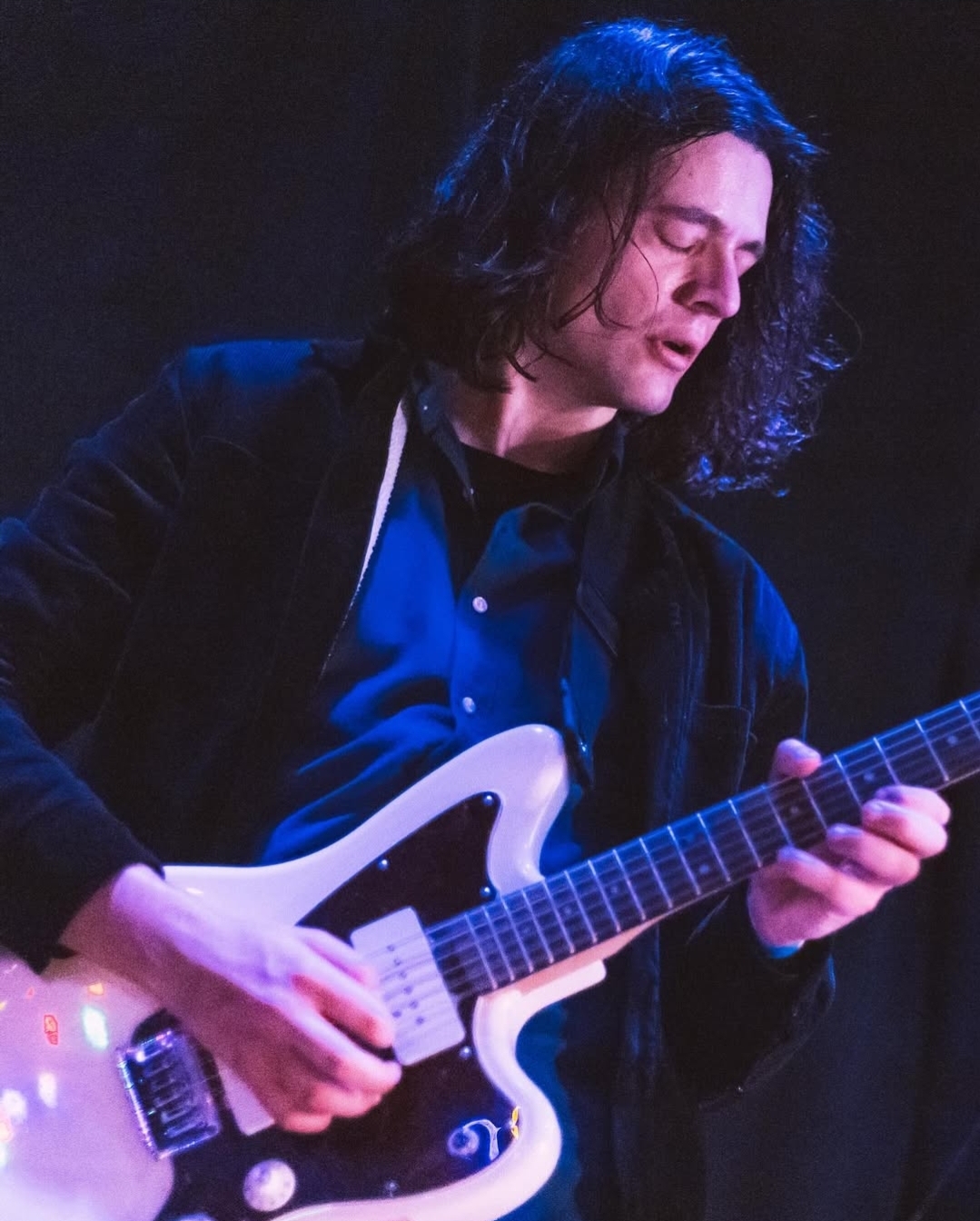 Person with long dark hair playing an electric guitar on stage, wearing a dark jacket and blue shirt.