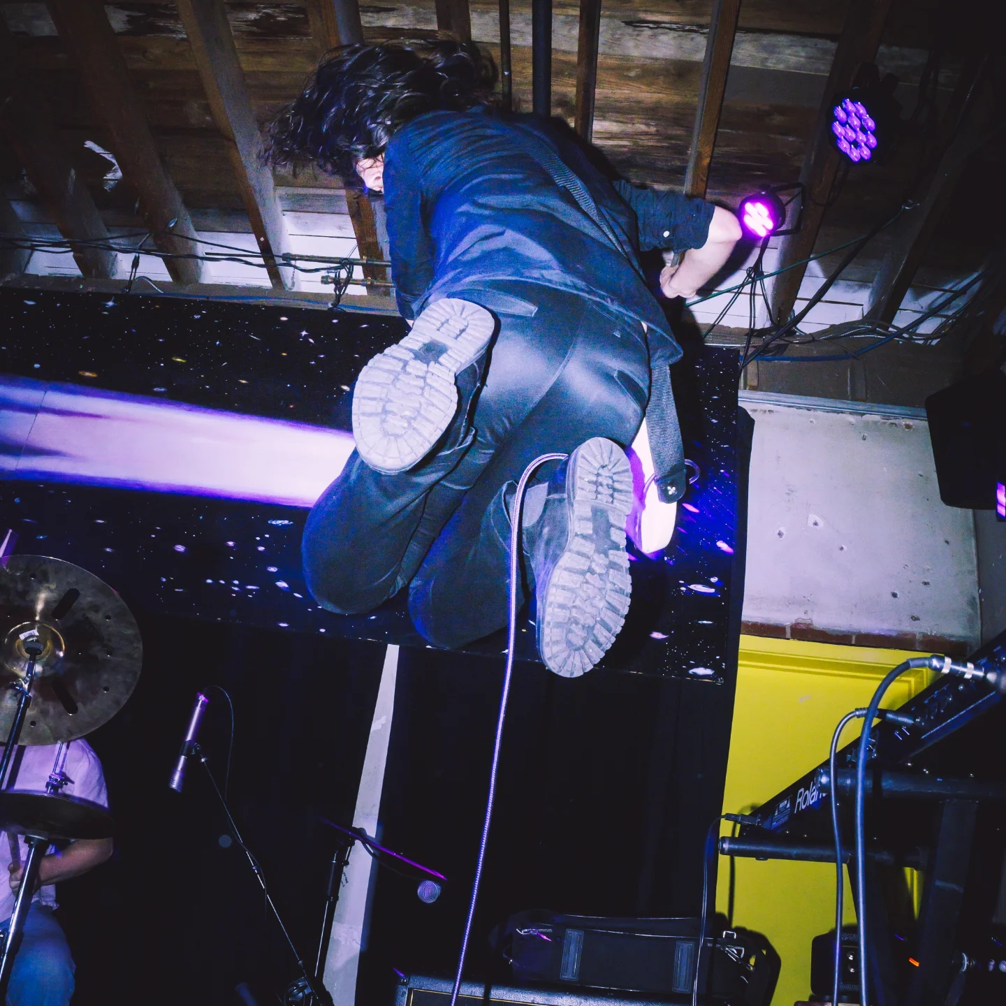 Person climbing a wall with purple lighting, ceiling beams visible above, wearing dark clothing and sneakers.
