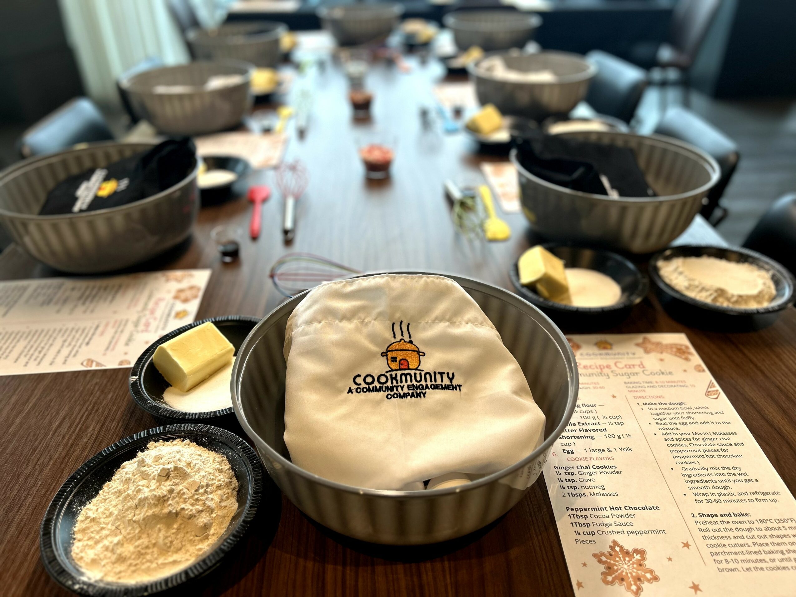 A dining table set with bowls, utensils, and ingredients for a cooking class, with a cloth in a bowl in the center.