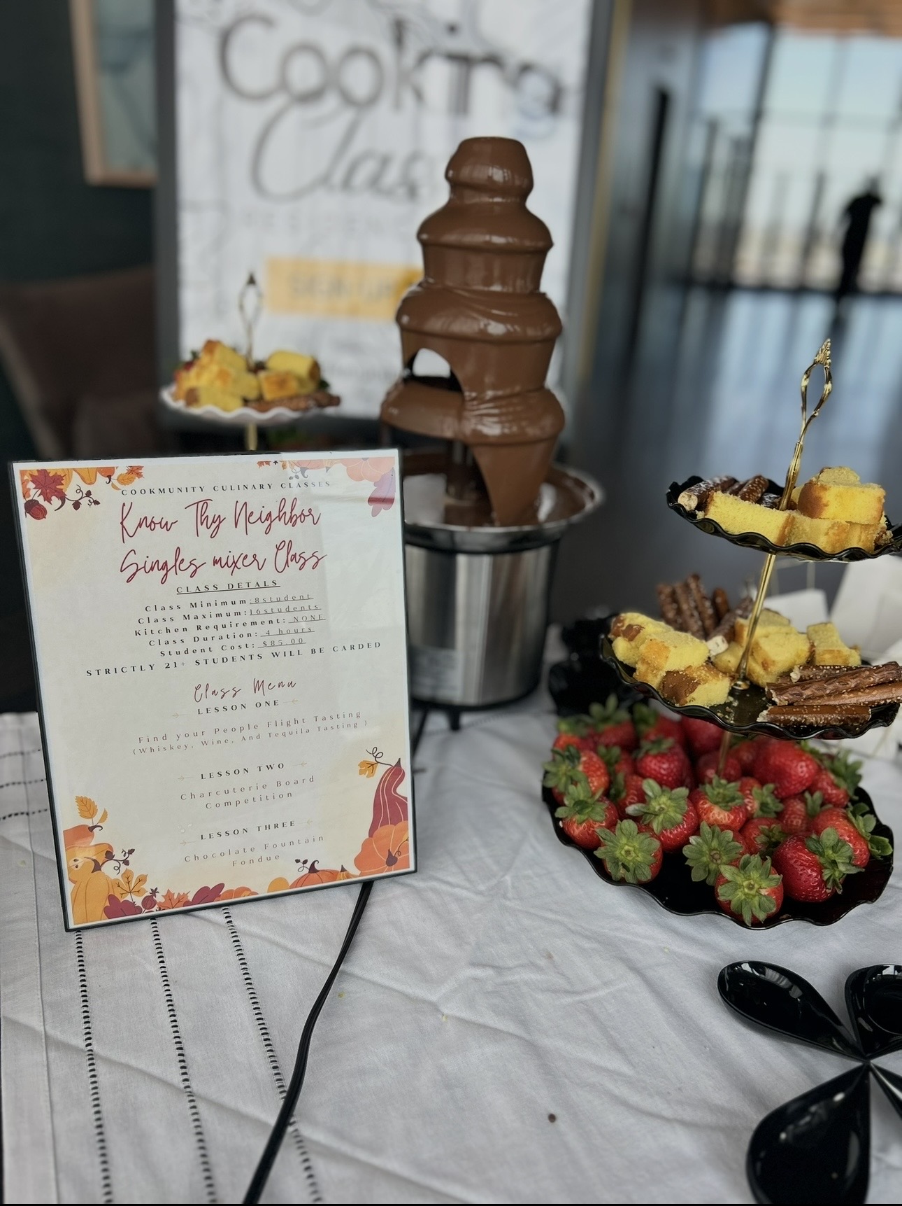 Chocolate fountain with strawberries and desserts on table, menu card nearby, indoor setting with window in background.