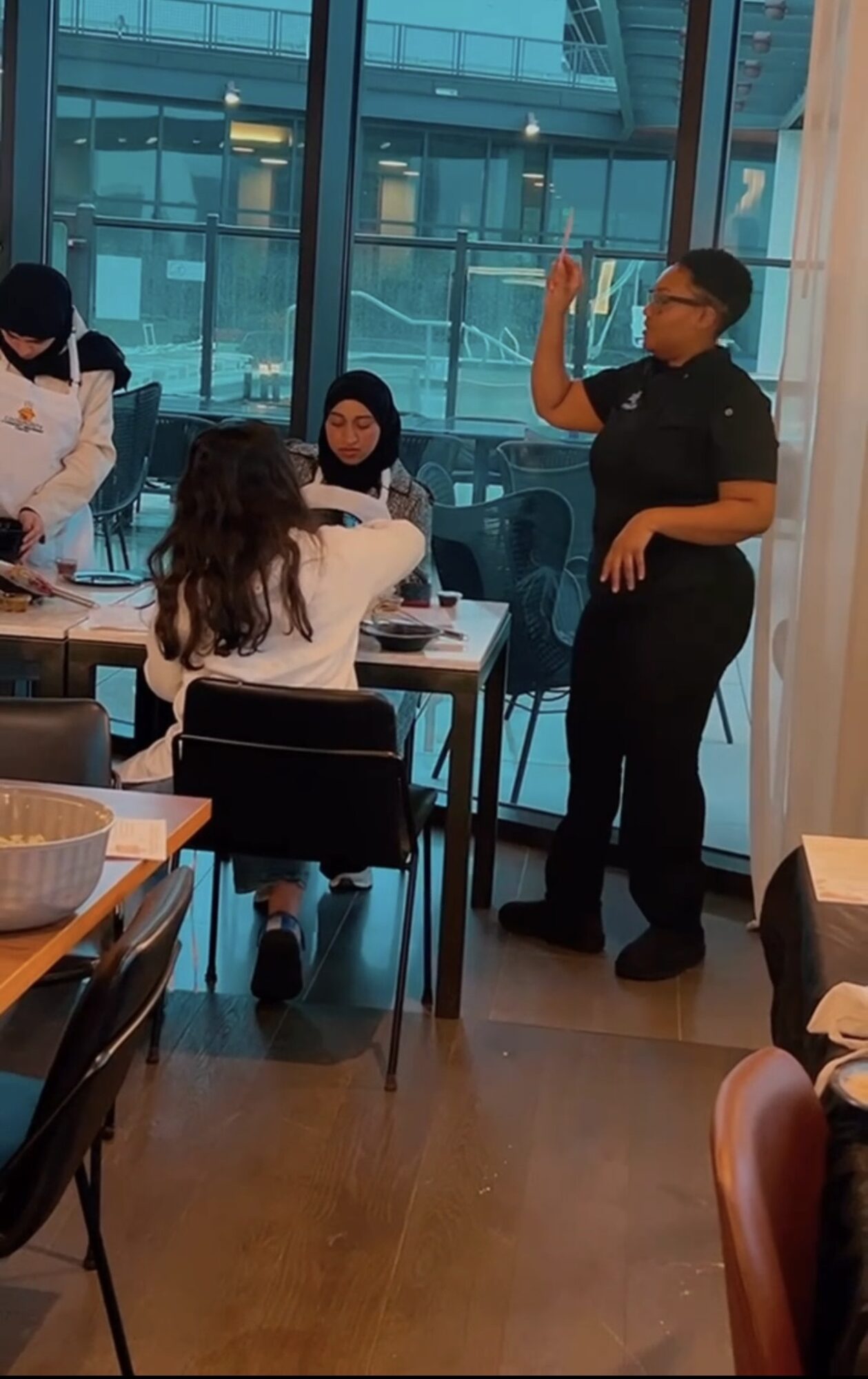 Group of people in a restaurant, with a person standing and gesturing, others seated at tables, large window in background.