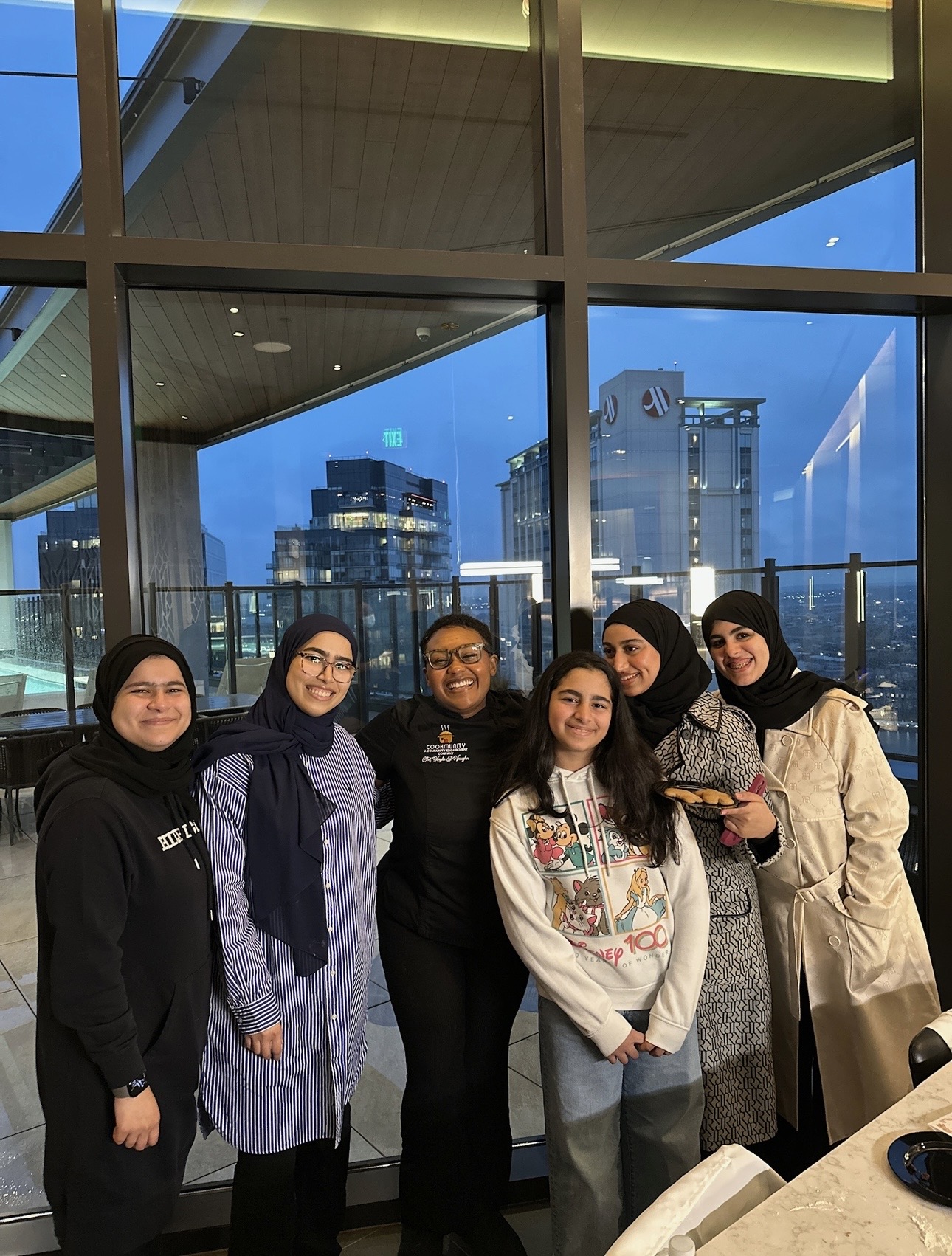 Six women standing together indoors with a cityscape view through large windows behind them, smiling and posing for the photo.