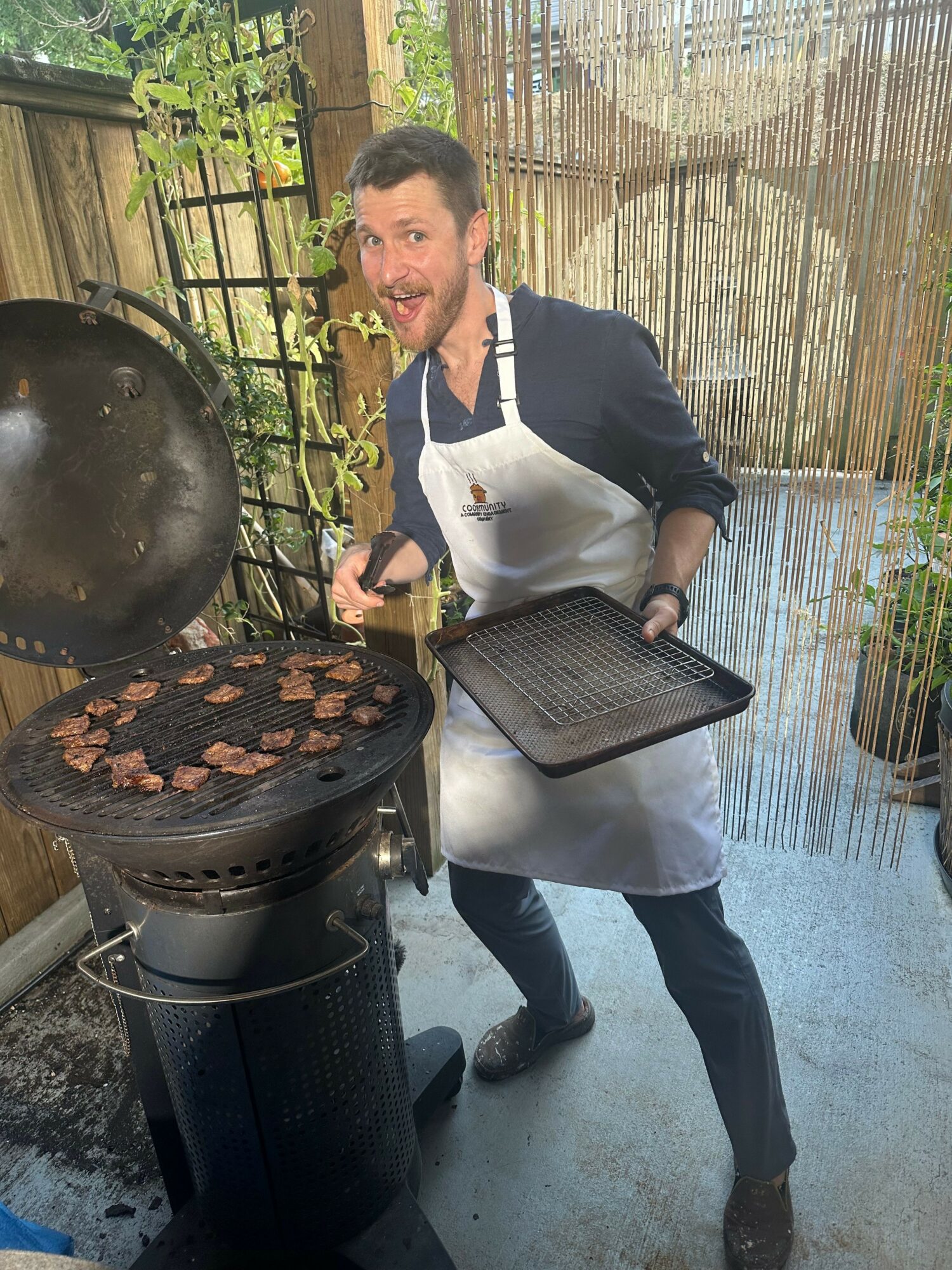 Man grilling meat outdoors, holding a tray, with a barbecue grill in front, bamboo fence in background, daytime.
