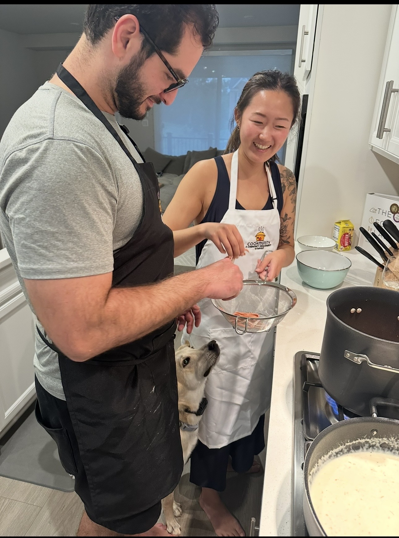 Two people cooking in a kitchen, smiling, with a small dog looking up at them, surrounded by cooking utensils and ingredients.